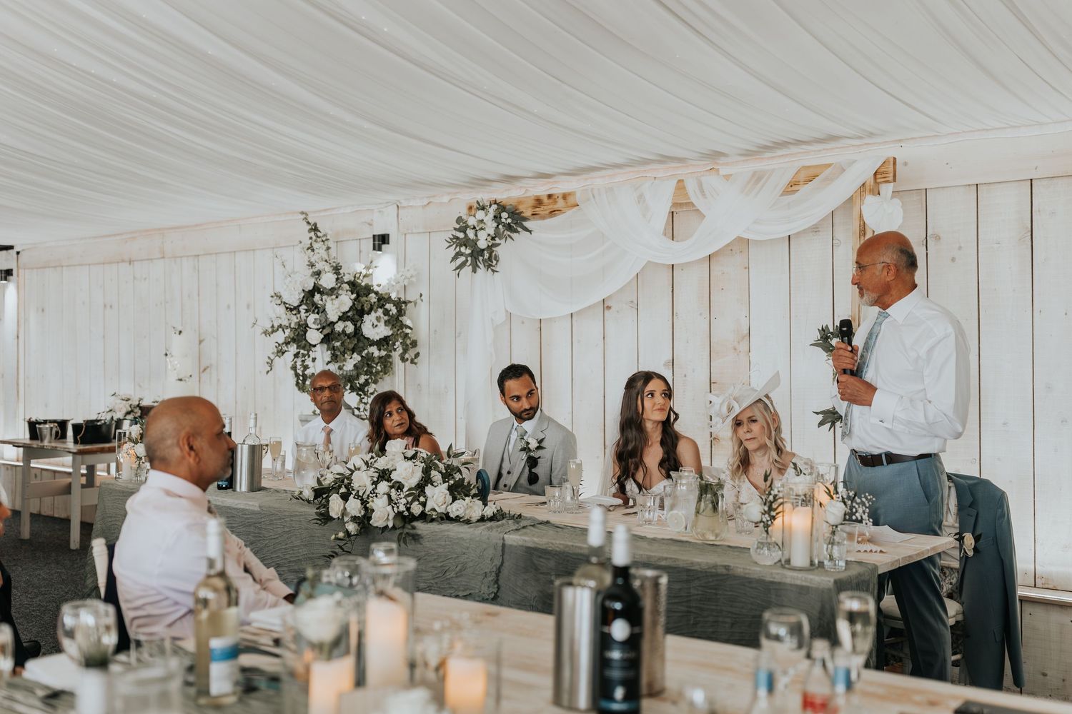 Wedding reception speech by guest at elegant white marquee venue with floral arrangements and bridal party seated at head table.