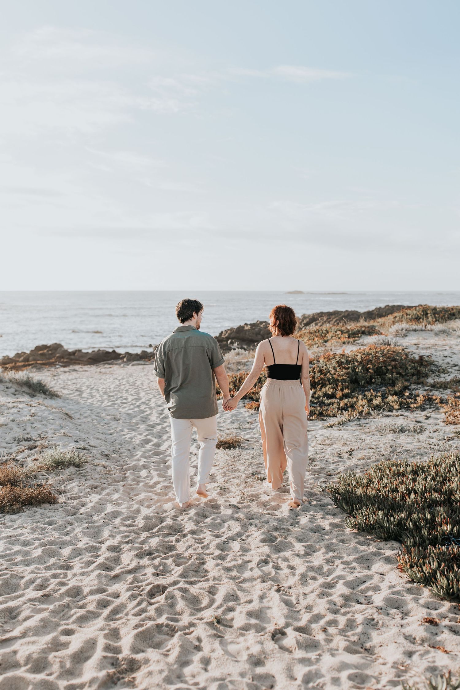 A couple walking together on a sandy beach with rocky outcroppings and the ocean in the background.