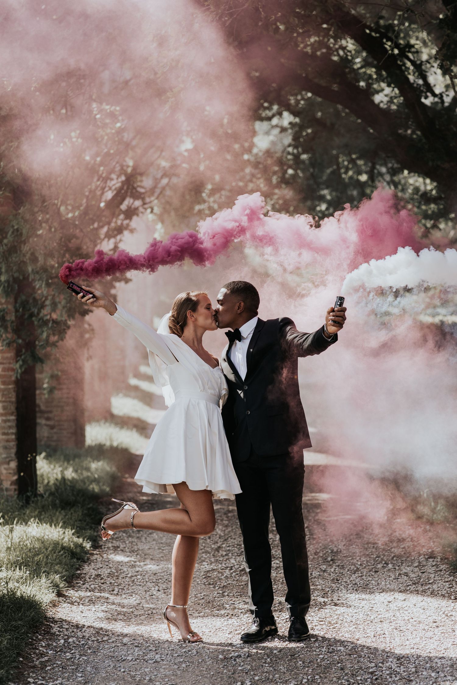 A bride and groom share a romantic moment amidst colorful smoke in a scenic outdoor setting.