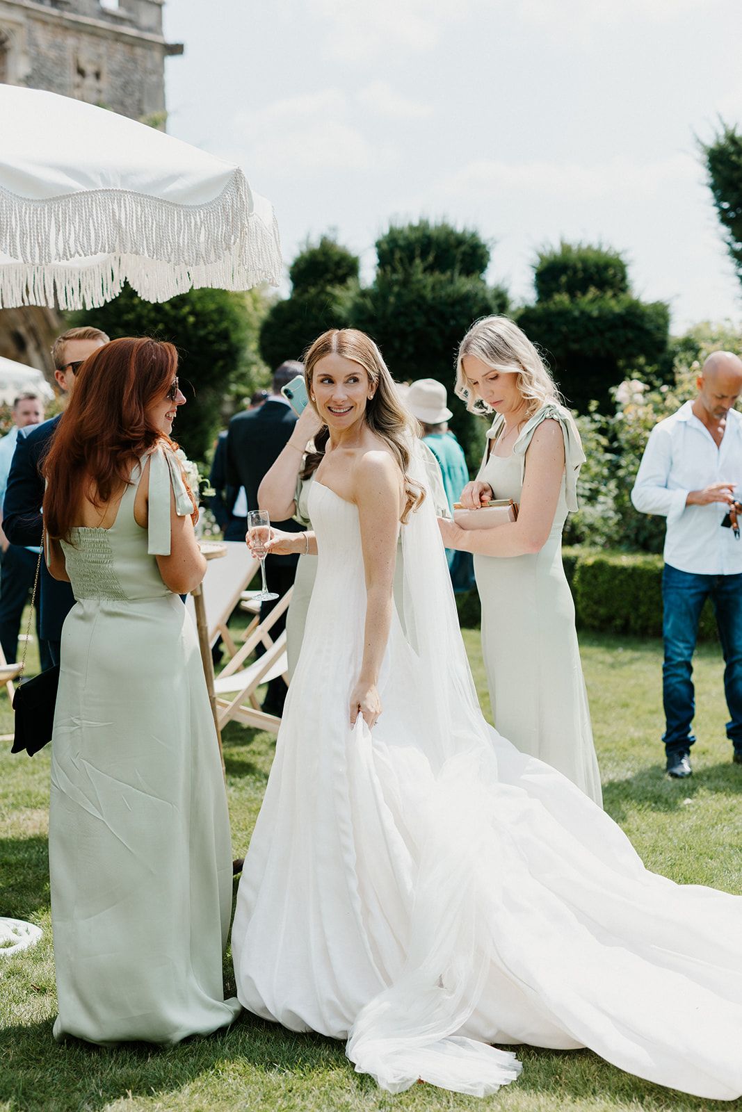 Bride in white gown smiles with bridesmaids in sage green dresses at outdoor wedding reception.