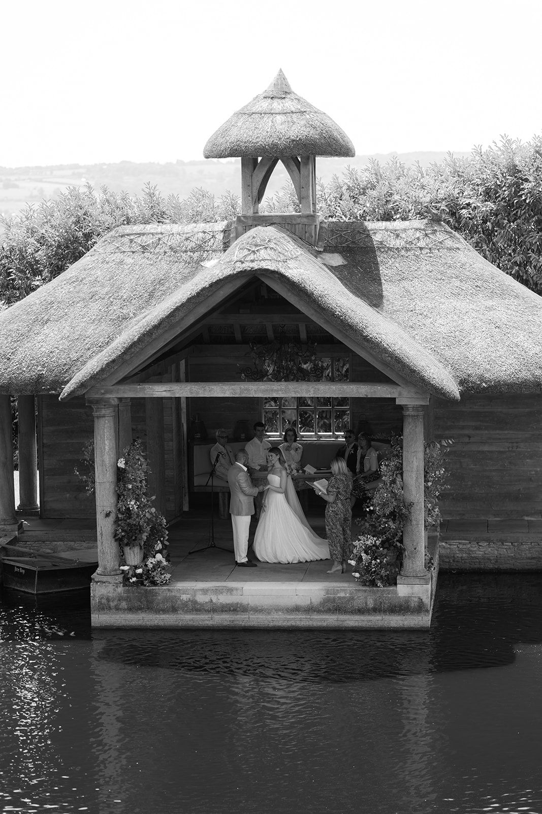 Black and white photo of a wedding ceremony in a thatched boathouse over water.