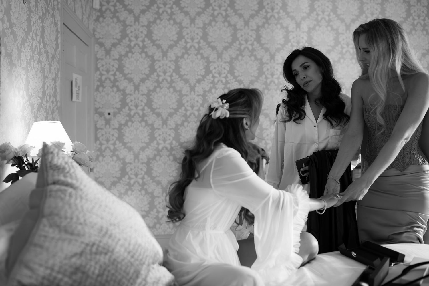 Black and white photo of a bride in white gown with floral headpiece being assisted by two women in a elegant bedroom.