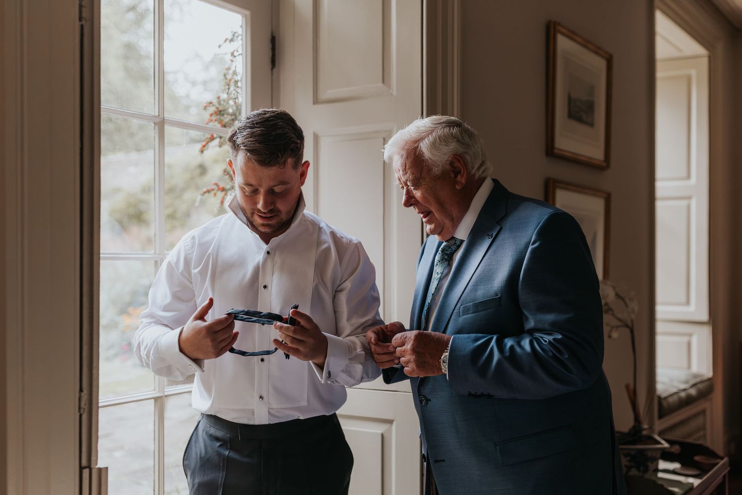 Two men in formal attire examining a bow tie near a bright window, preparing for a wedding celebration.