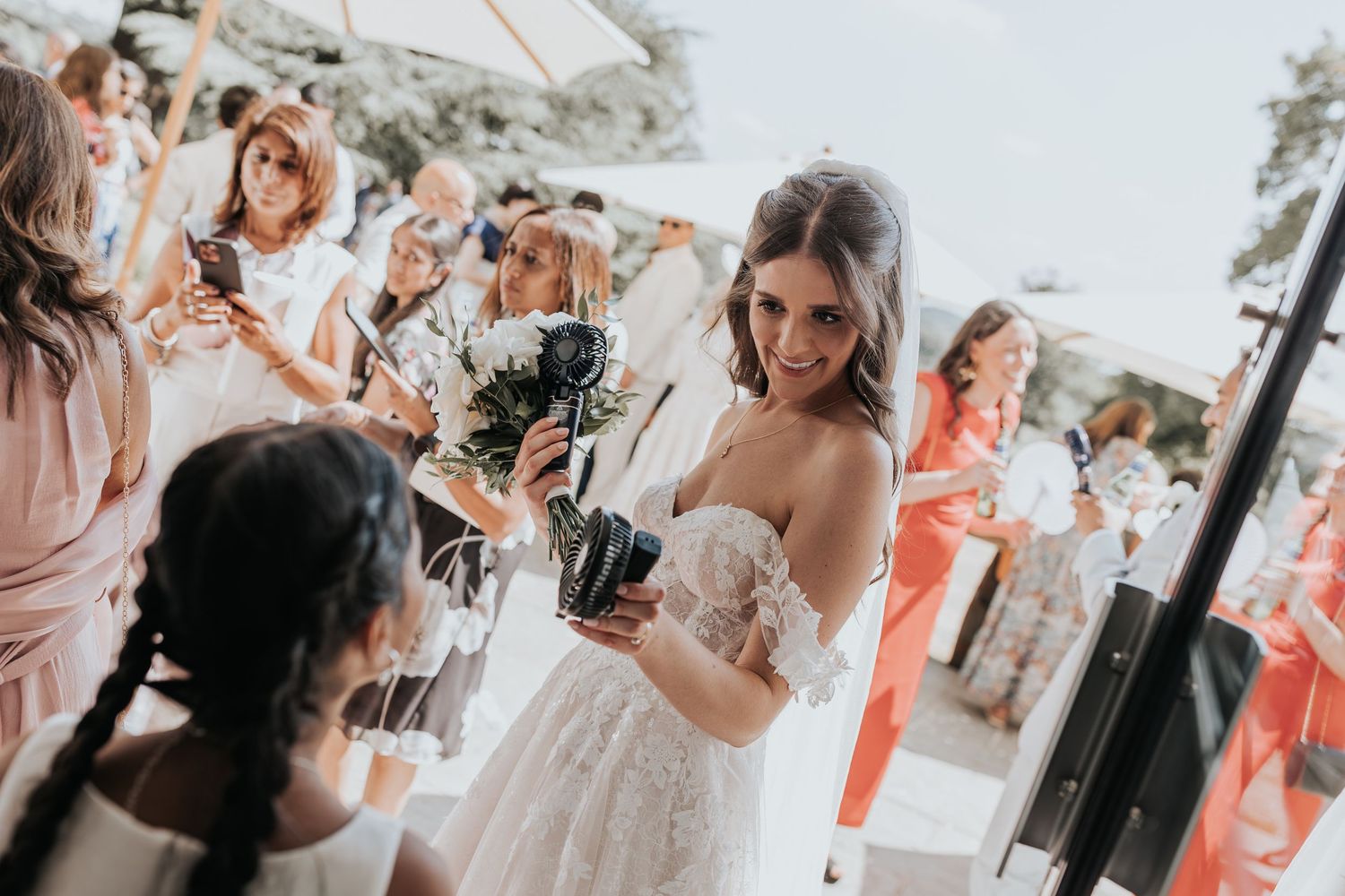 Bride in lace wedding dress smiling while holding bouquet surrounded by guests at outdoor wedding celebration.