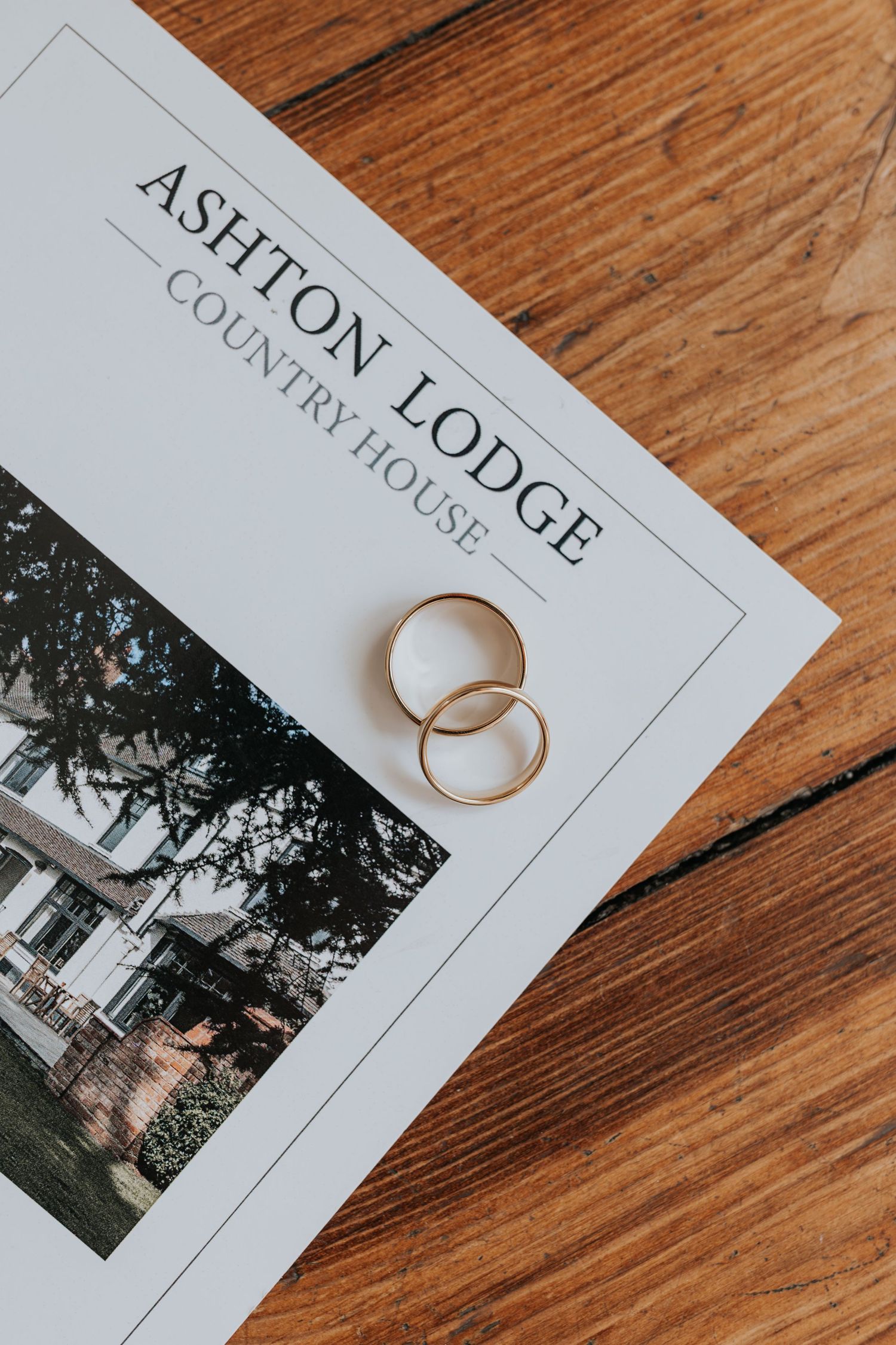 Two gold wedding rings resting on an Ashton Lodge Country House brochure on a wooden surface.