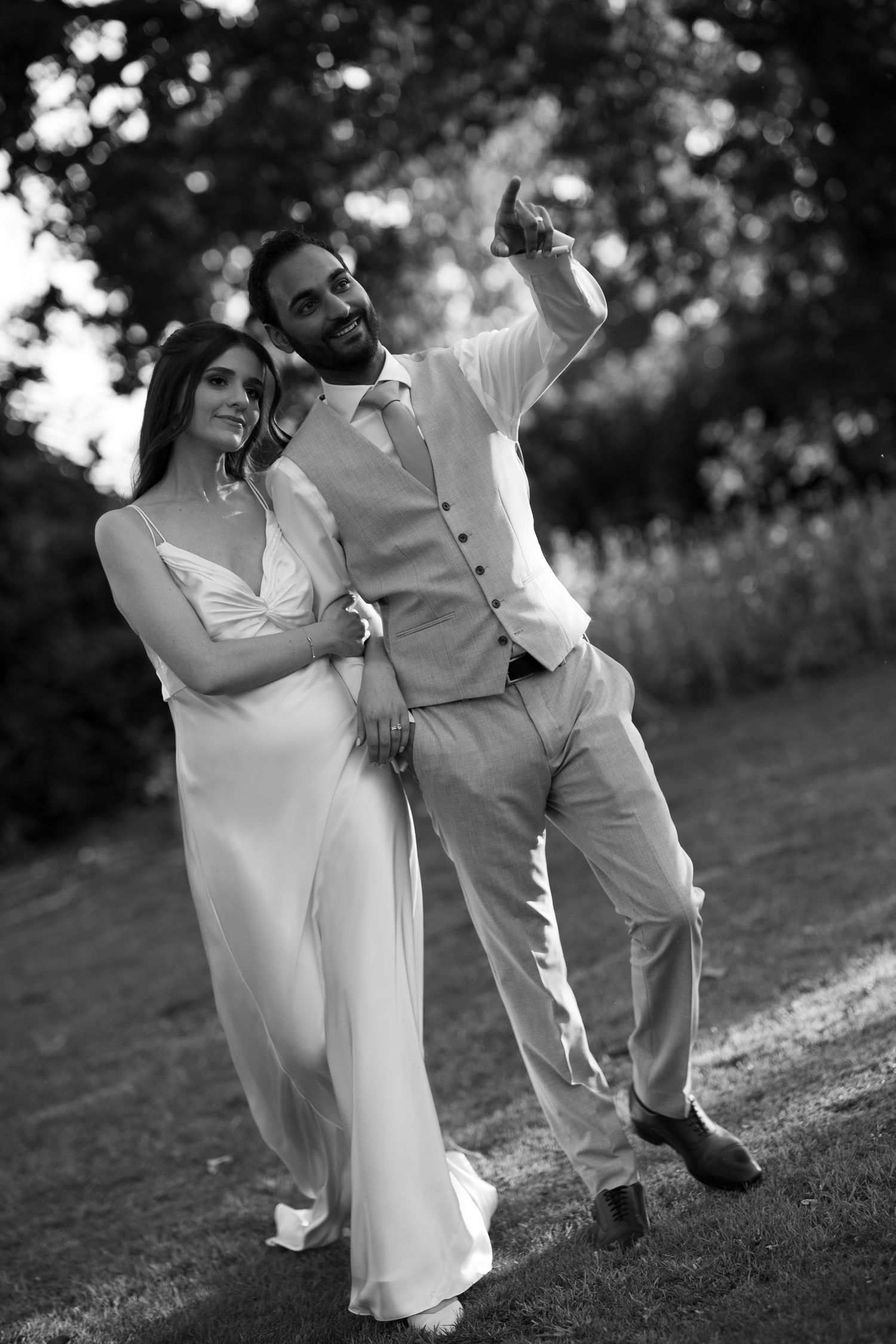 Black and white photo of a happy couple in wedding attire outdoors, groom pointing upward while bride smiles beside him.