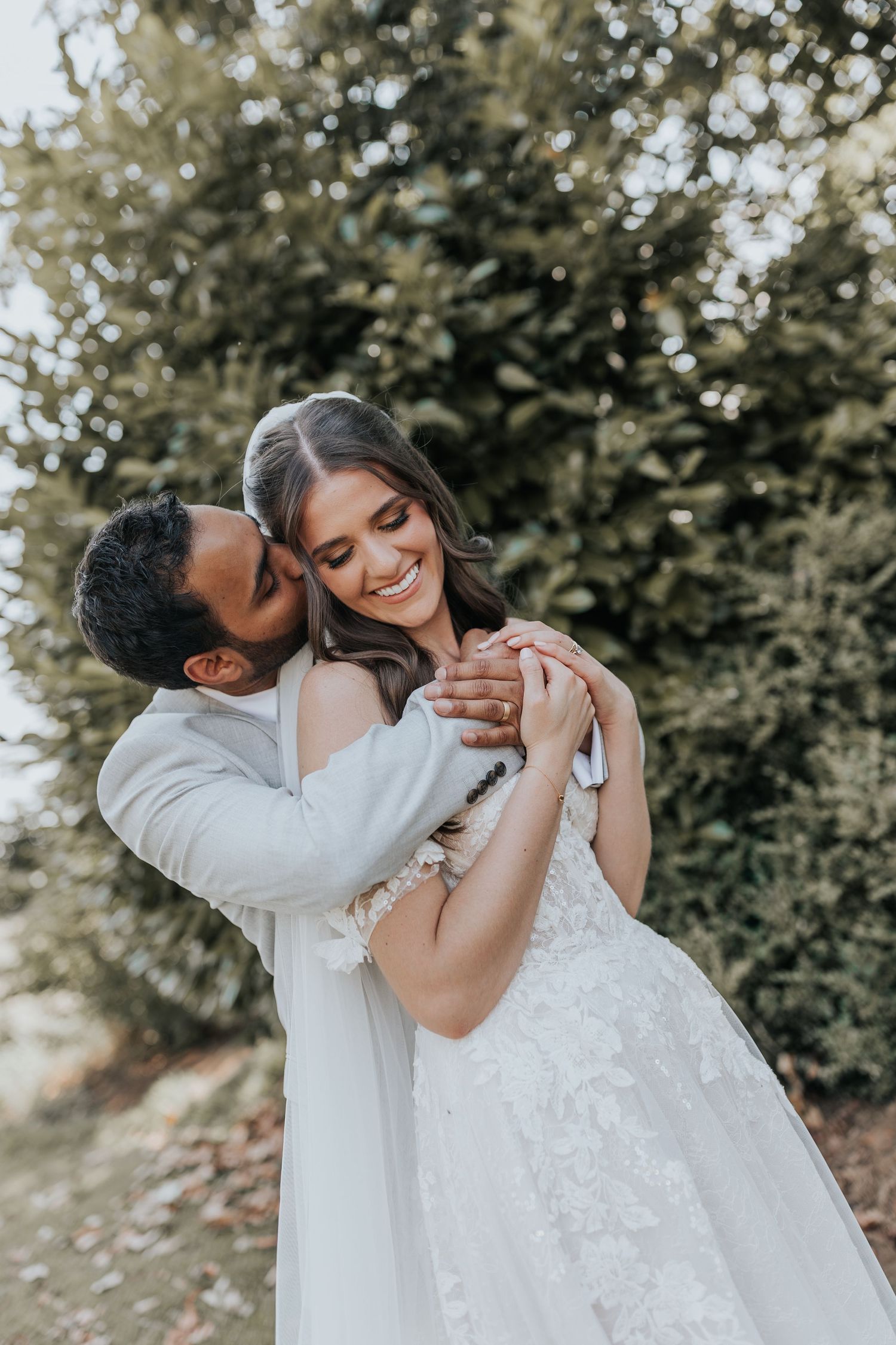 Groom in white suit kisses bride on cheek while holding hands outdoors among lush green trees.
