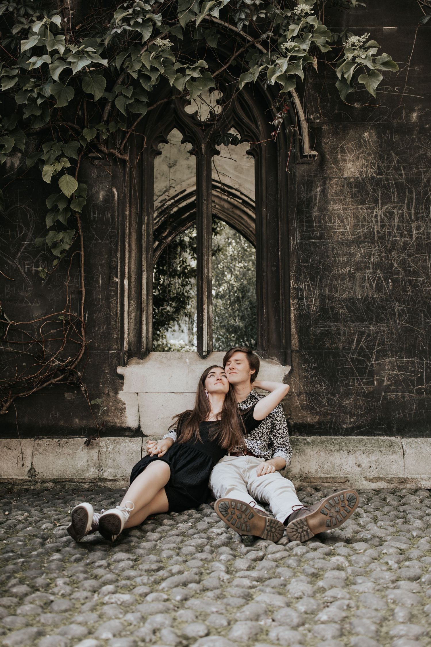 Two young women sitting on the ground in front of an ornate stone archway surrounded by ivy-covered walls.