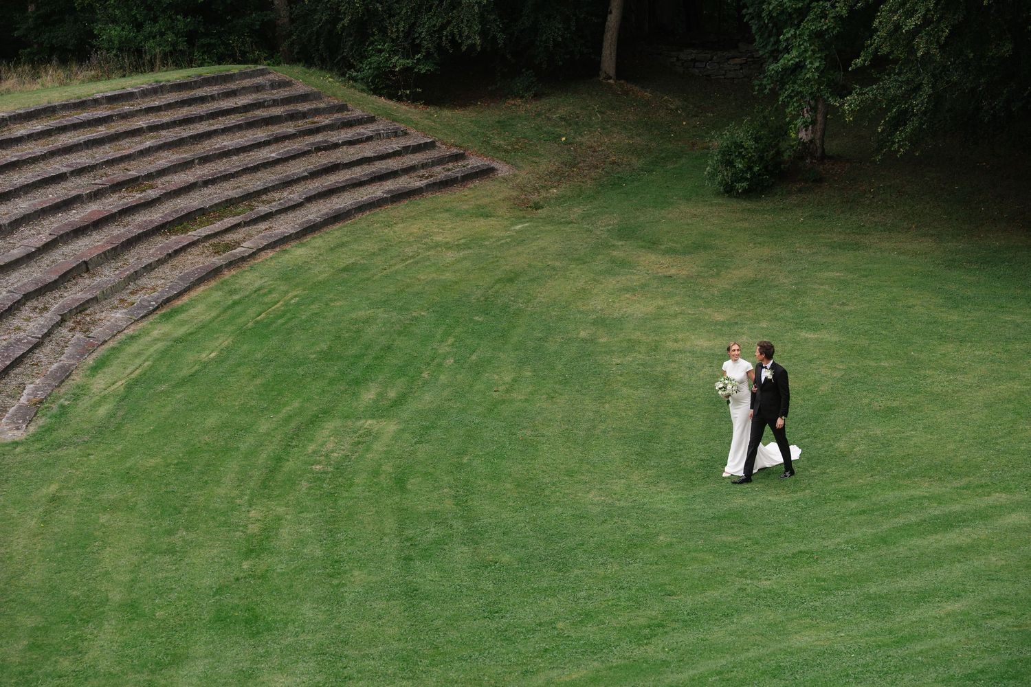Aerial view of a bride and groom walking across a lush green lawn near curved stone steps at their venue.