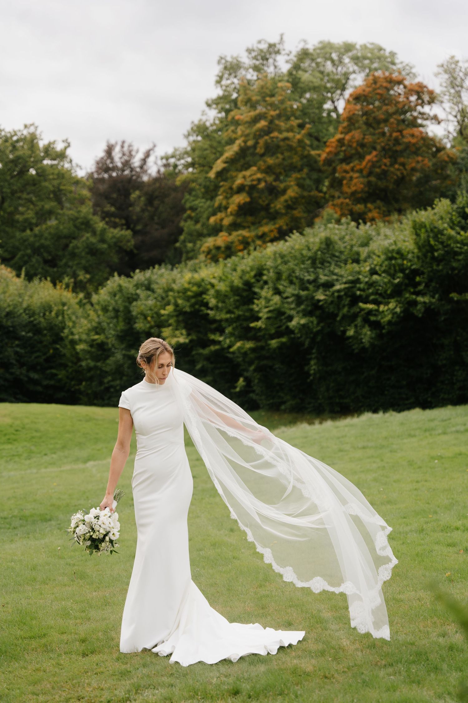 Elegant bride in fitted white gown with flowing lace-edged veil billowing in garden breeze.