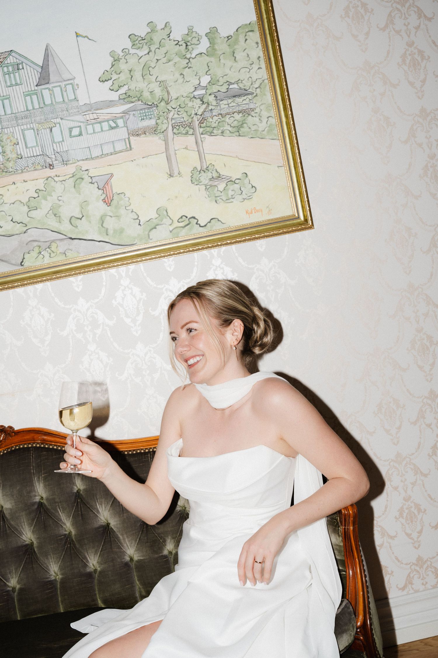 Smiling bride in white dress holds wine glass seated on vintage sofa indoors.