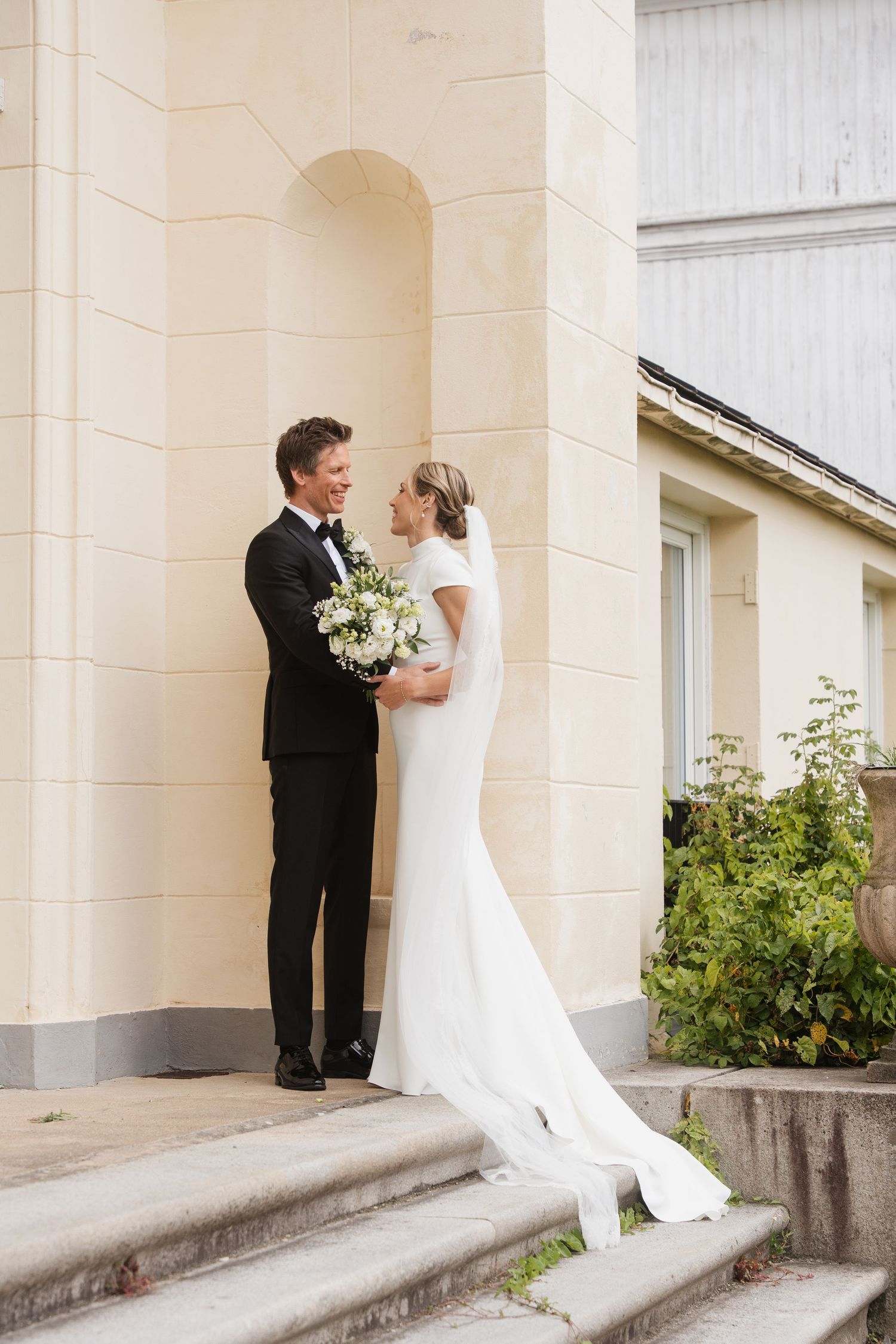 Bride and groom share a romantic moment on stone steps beside a cream-colored building after their wedding.