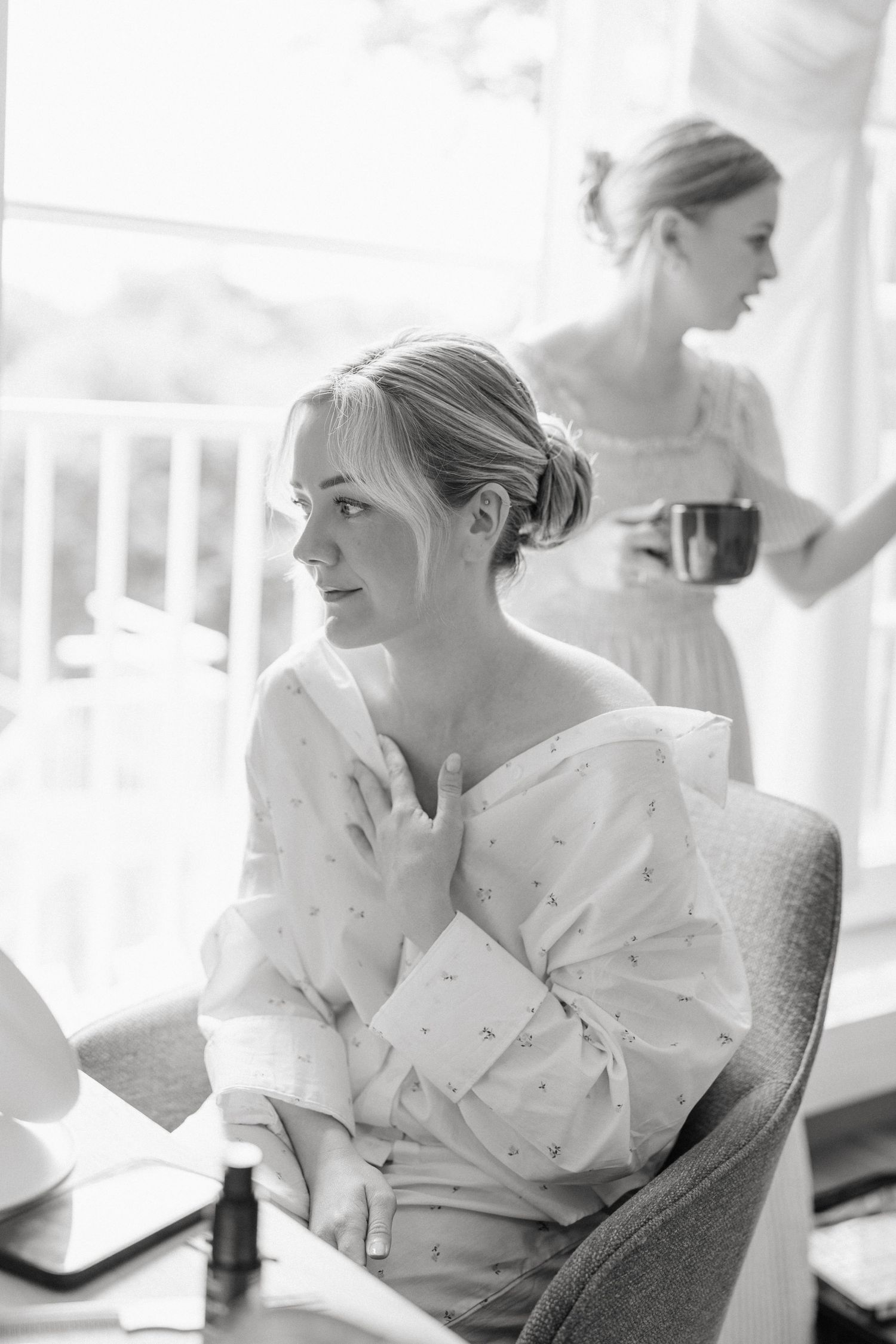Black and white photo of a bride in a floral robe sitting in a chair while getting ready on her wedding day.