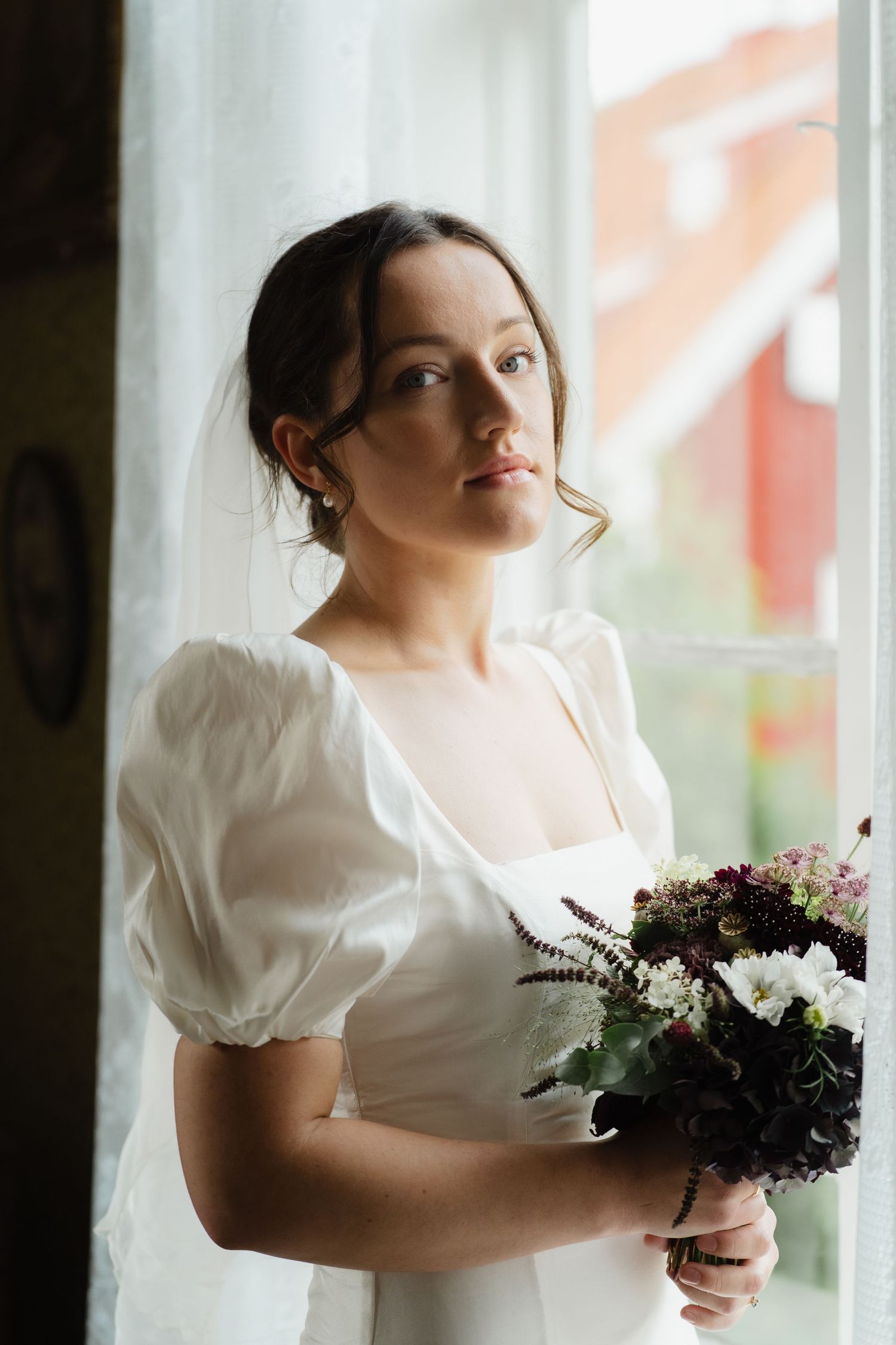 Serene bride in a white puff-sleeve wedding dress holding a dark floral bouquet while gazing out a window.