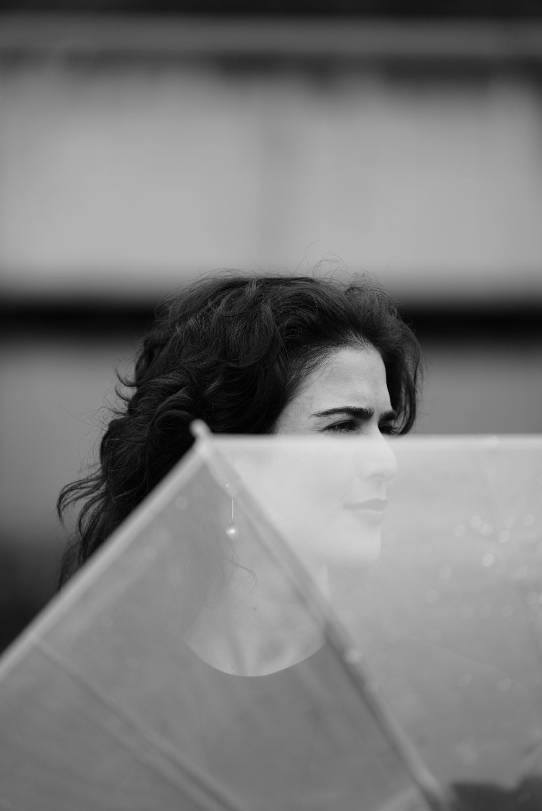 Black and white portrait of a woman with curly hair partially hidden behind a transparent umbrella in the rain.