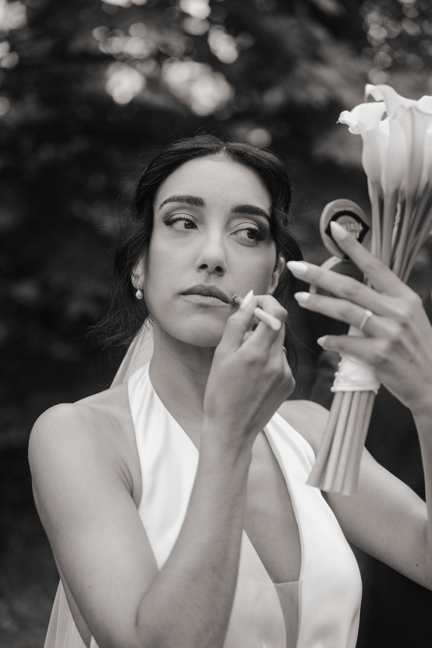 Black and white photo of a bride applying lipstick outdoors, holding a mirror and white floral bouquet.