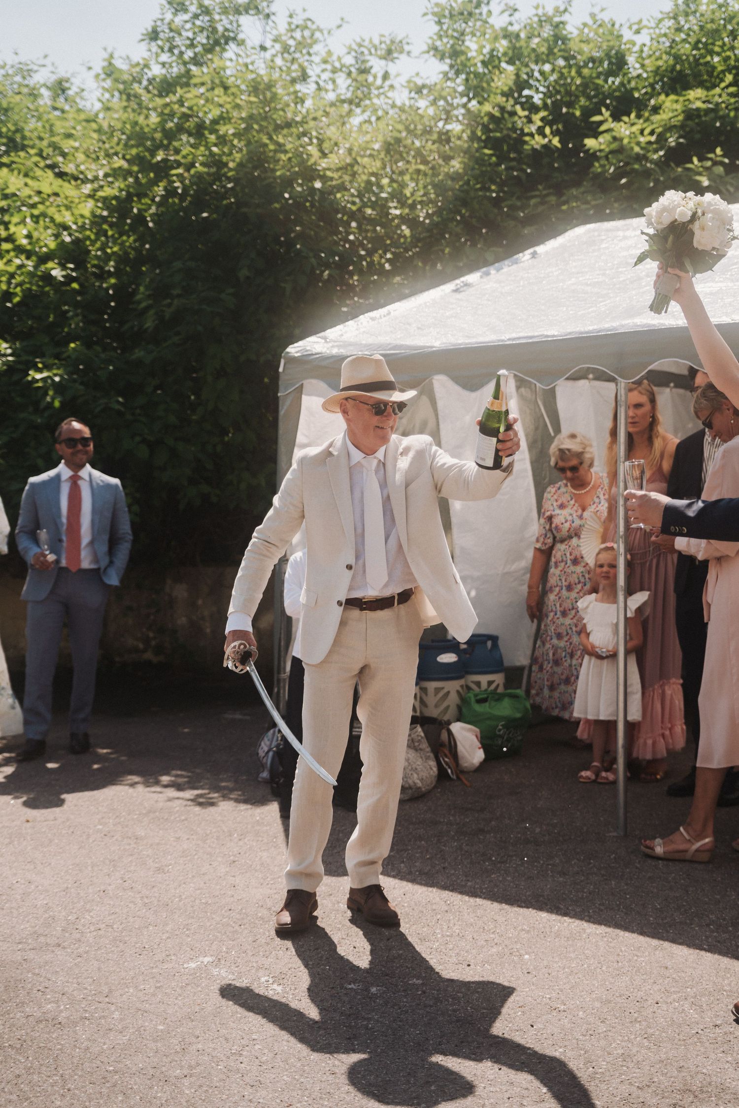 A man in a cream suit and hat celebrates at a wedding reception, holding a champagne bottle outdoors.