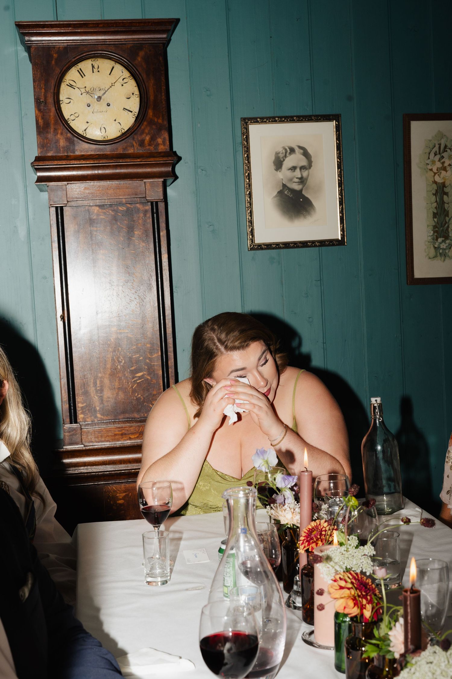 Emotional wedding guest crying at dinner table with grandfather clock and vintage portrait on teal wall.