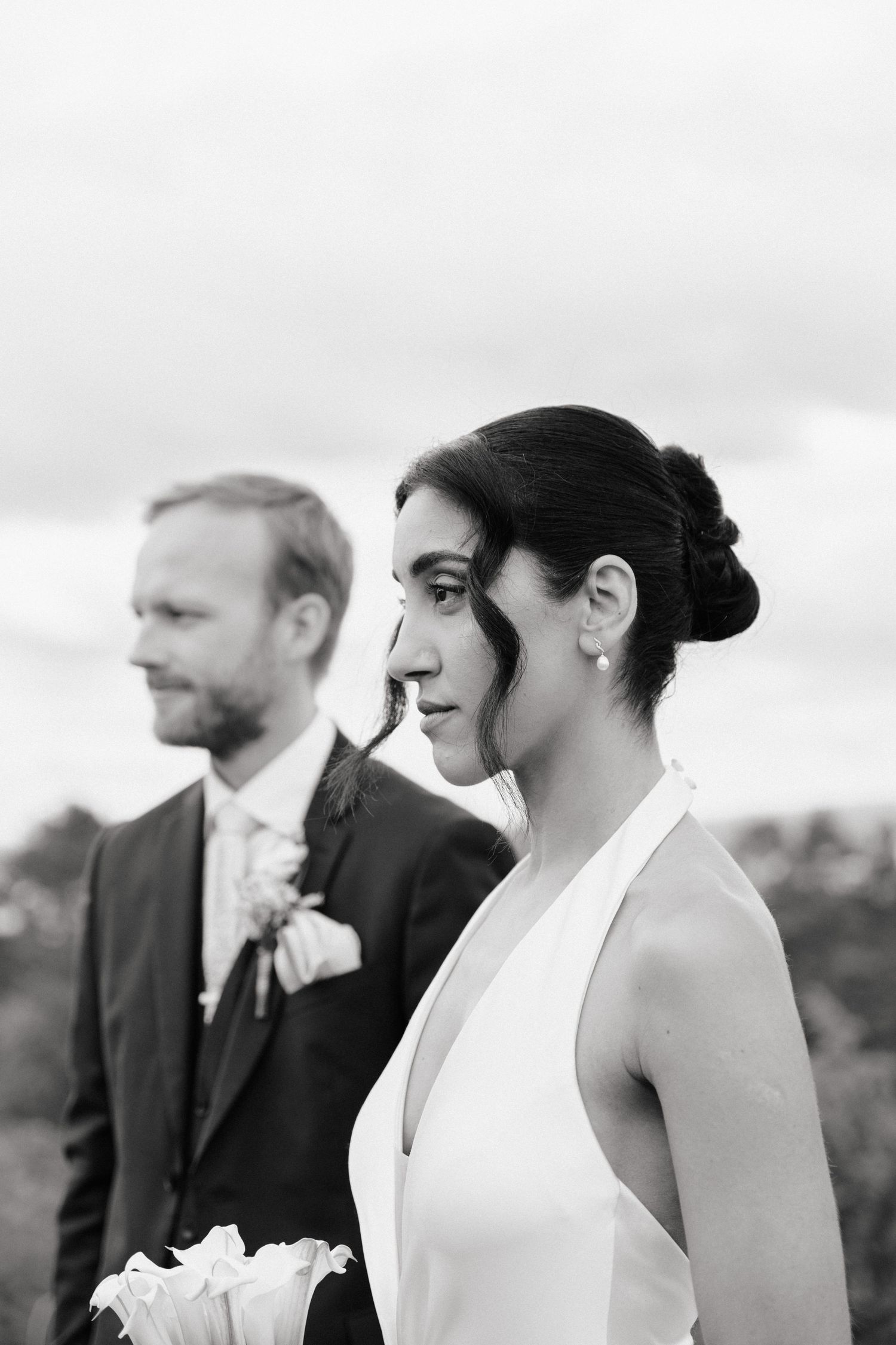 Black and white wedding photo of bride in white halter gown holding flowers with bearded groom in suit outdoors.