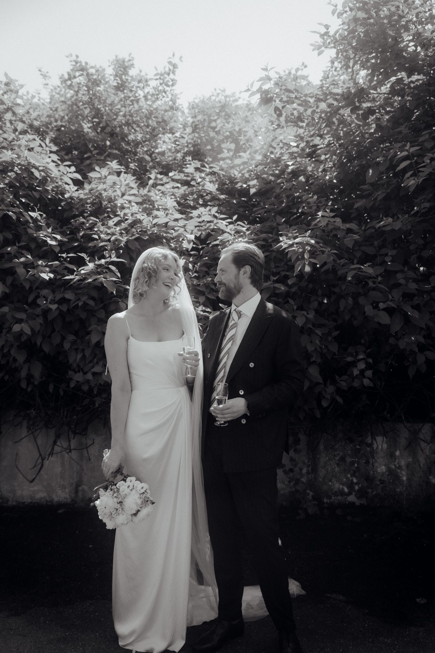 Black and white photo of a bride and groom sharing a tender moment outdoors surrounded by lush greenery.