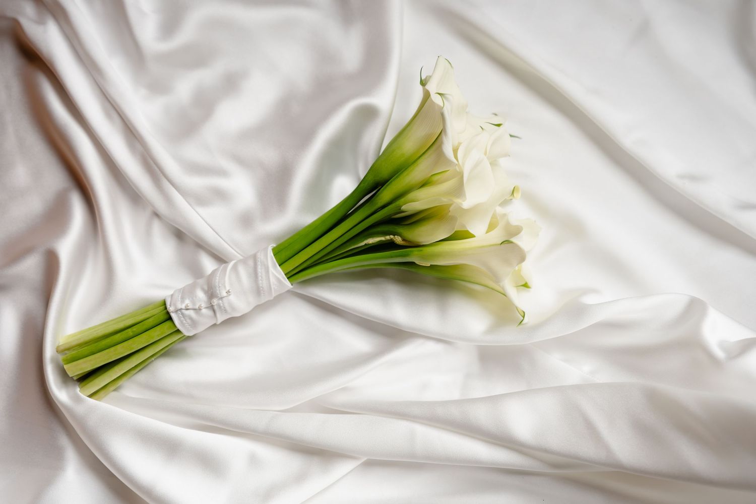 White calla lily bridal bouquet with lace ribbon resting on elegant white satin fabric.