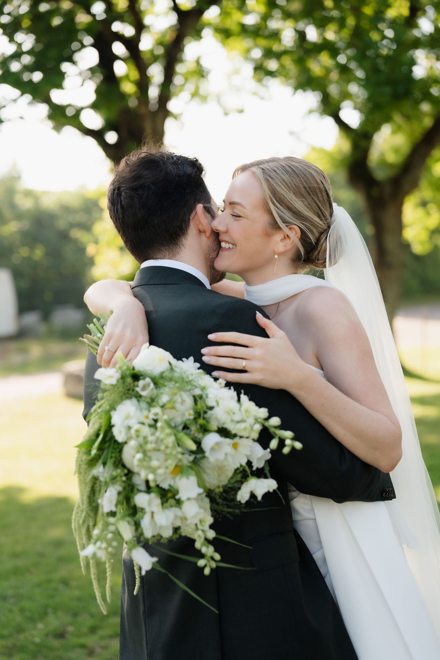 Happy bride in veil embraces groom holding white floral bouquet outdoors.