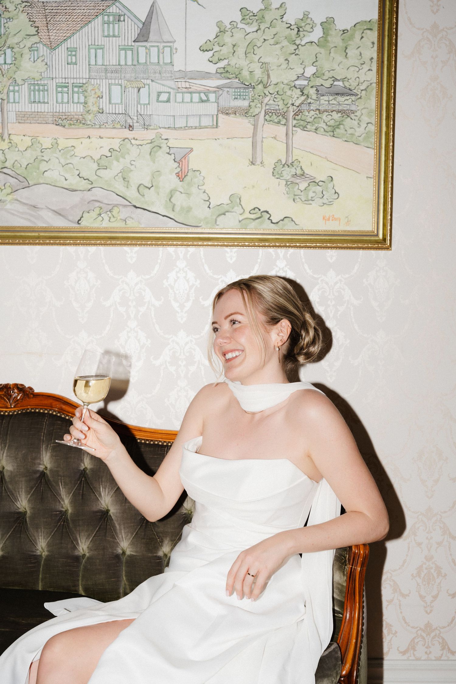 A smiling bride in a white halter gown sits on a vintage sofa holding a glass of white wine.