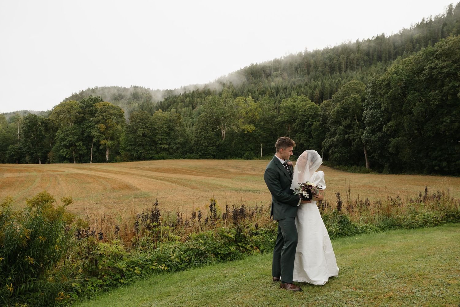Bride and groom share a romantic kiss in a scenic countryside field with misty forested hills.