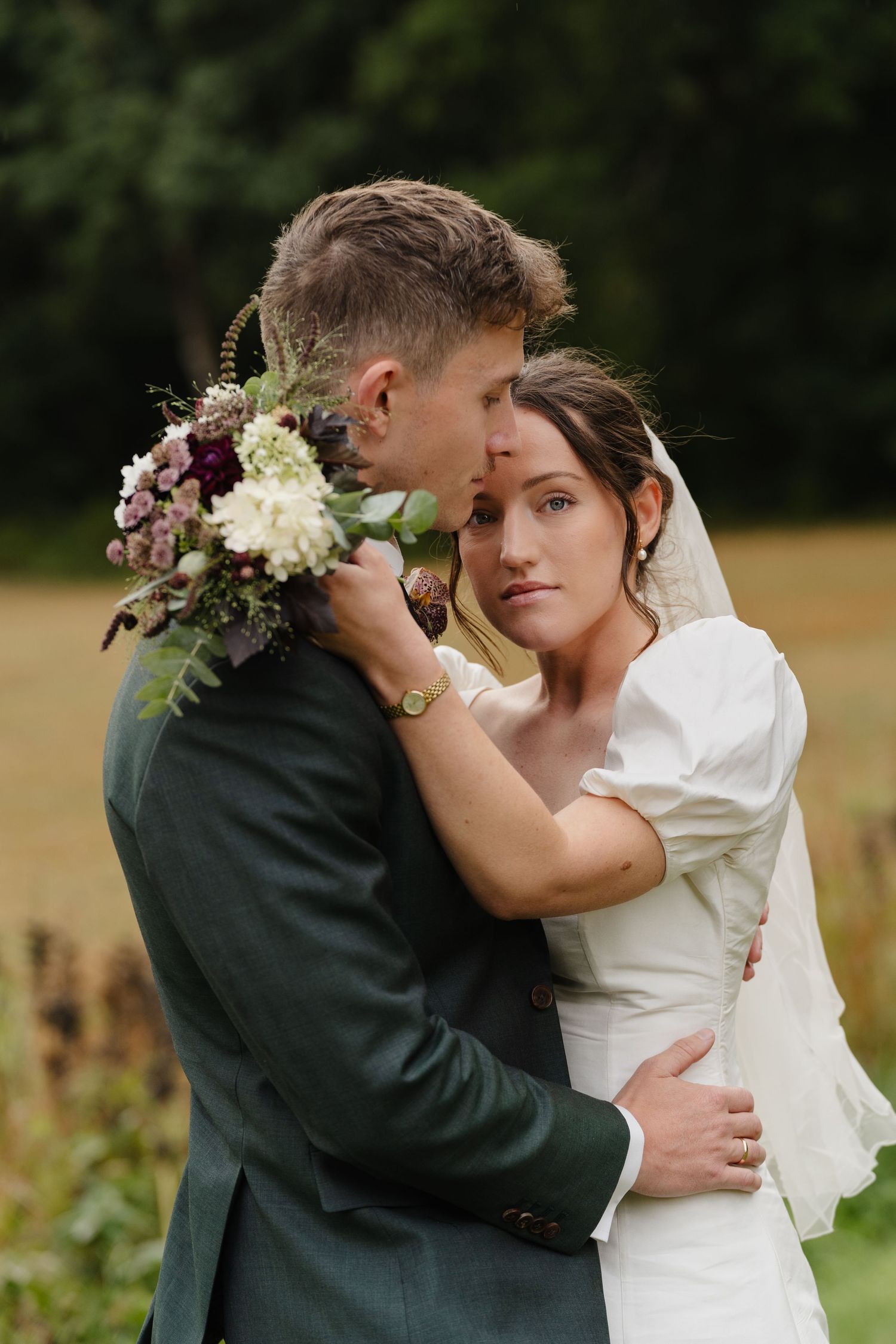 Bride holding bouquet of burgundy flowers while embracing groom in a green suit.