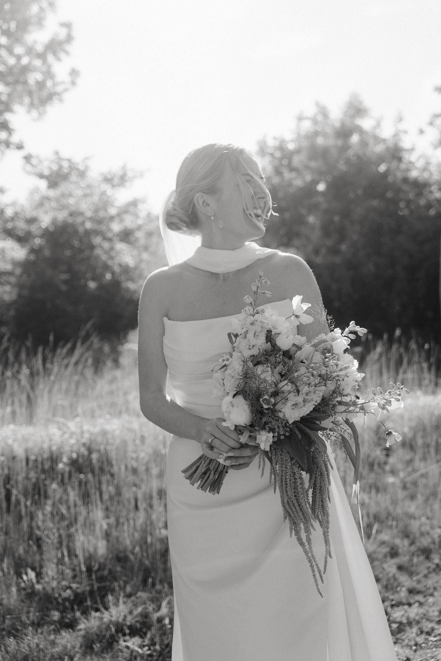 Black and white photo of a bride in a strapless gown holding a wildflower bouquet, standing in a sunlit field.