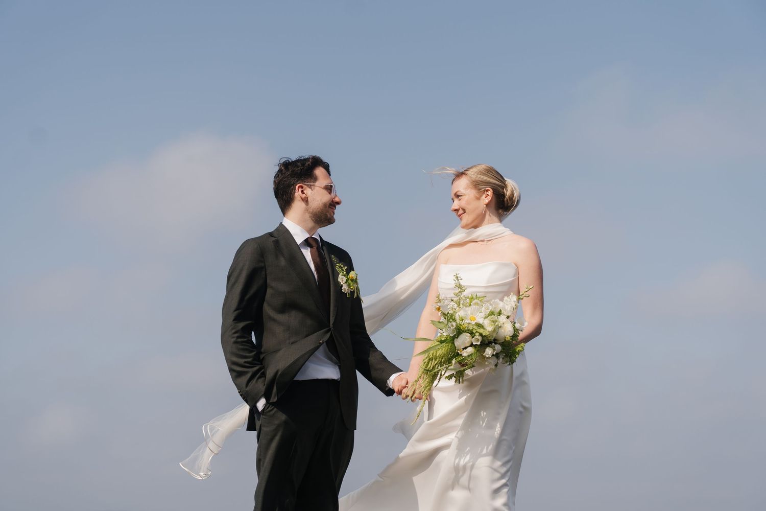 Bride and groom hold hands smiling outdoors with veil blowing in wind against blue sky.
