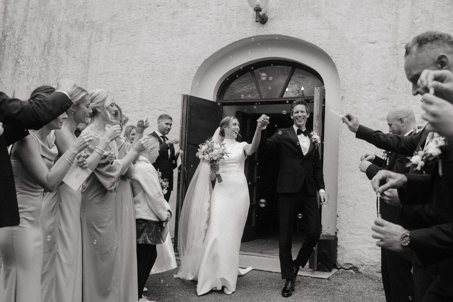 Black and white photo of a newlywed couple exiting a church as guests celebrate with confetti.