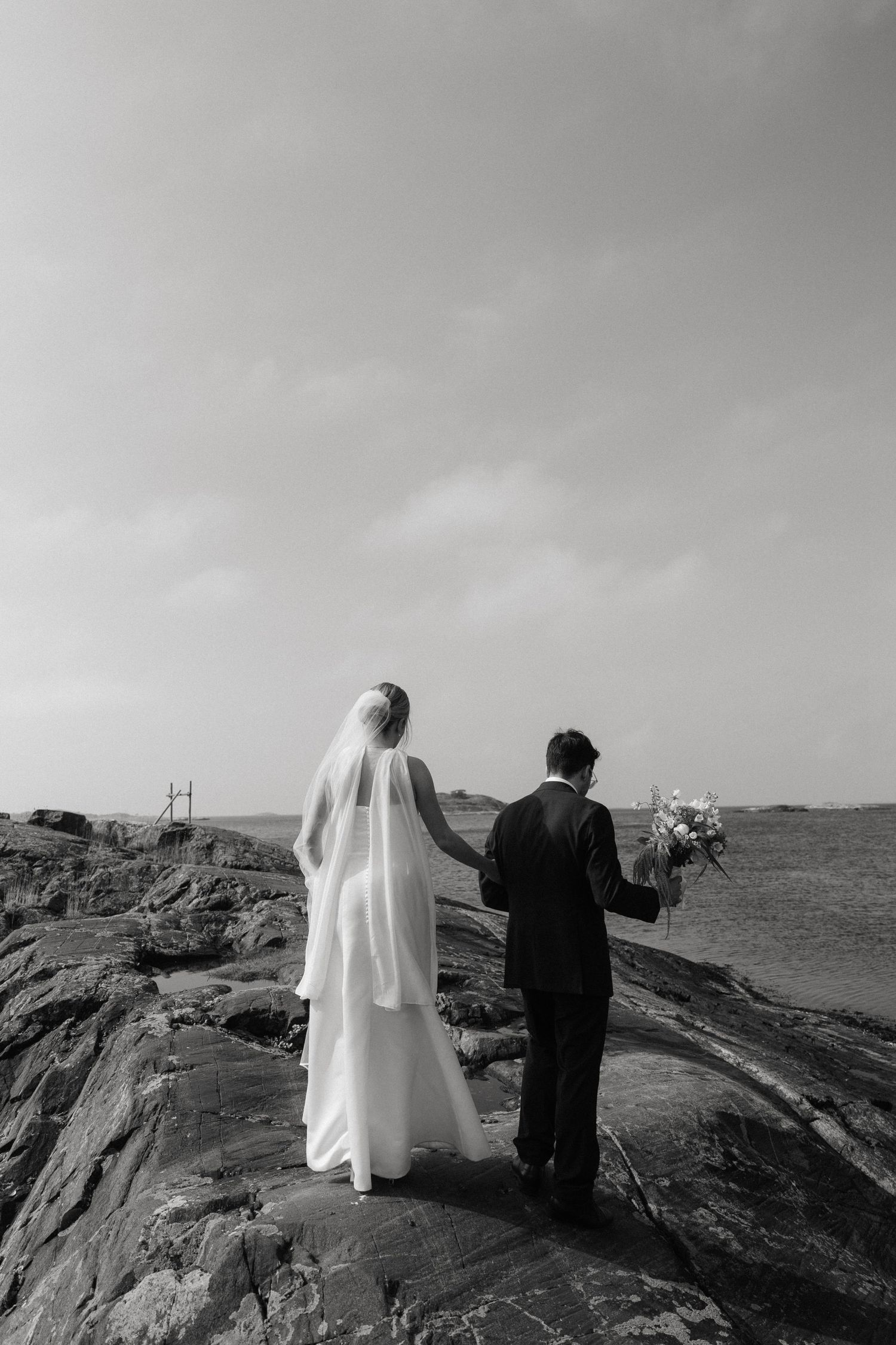 Black and white photo of bride and groom holding hands on rocky coastal landscape, bride in veil holding bouquet.
