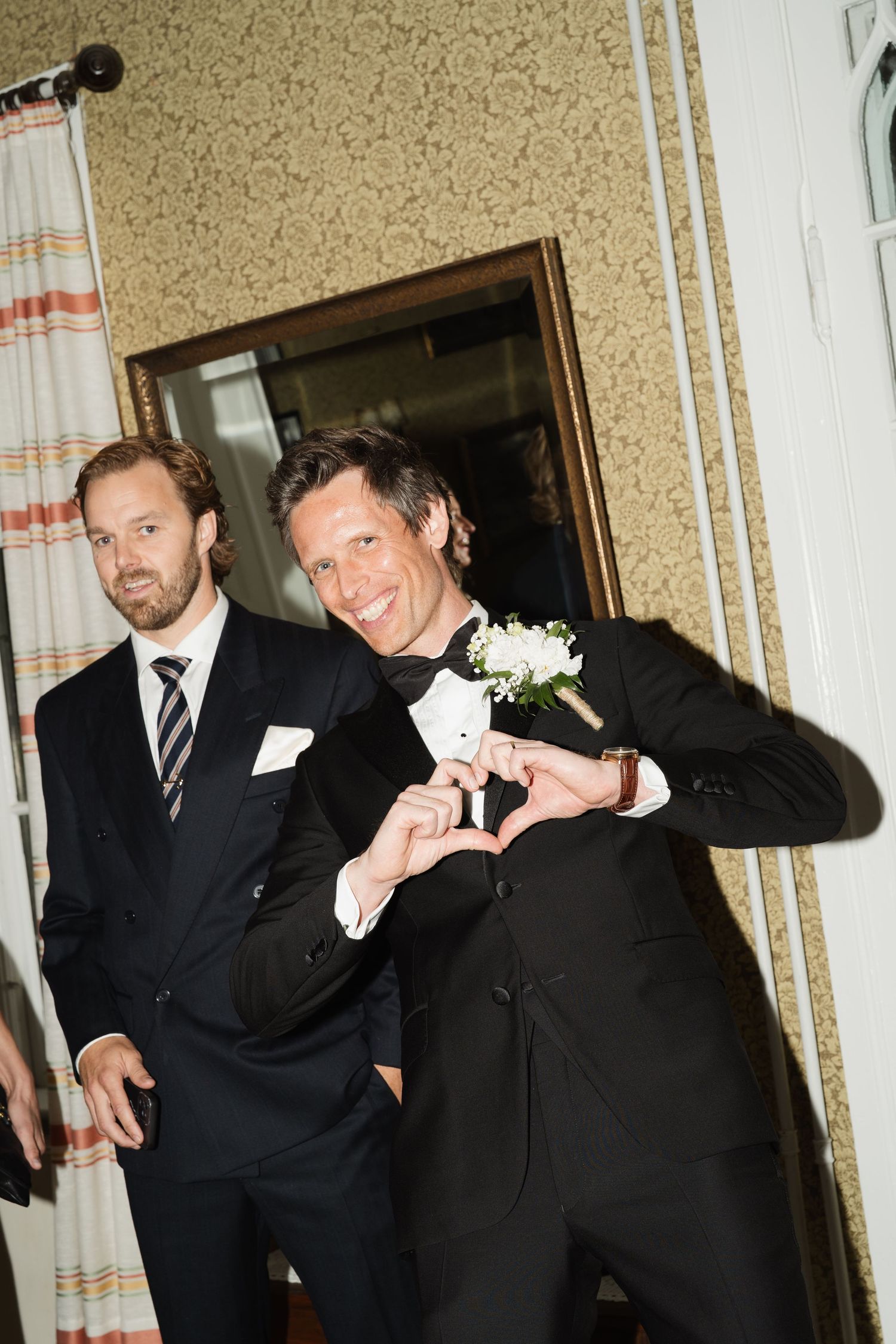 Smiling groom in tuxedo makes a heart shape with his hands while a groomsman stands behind him.