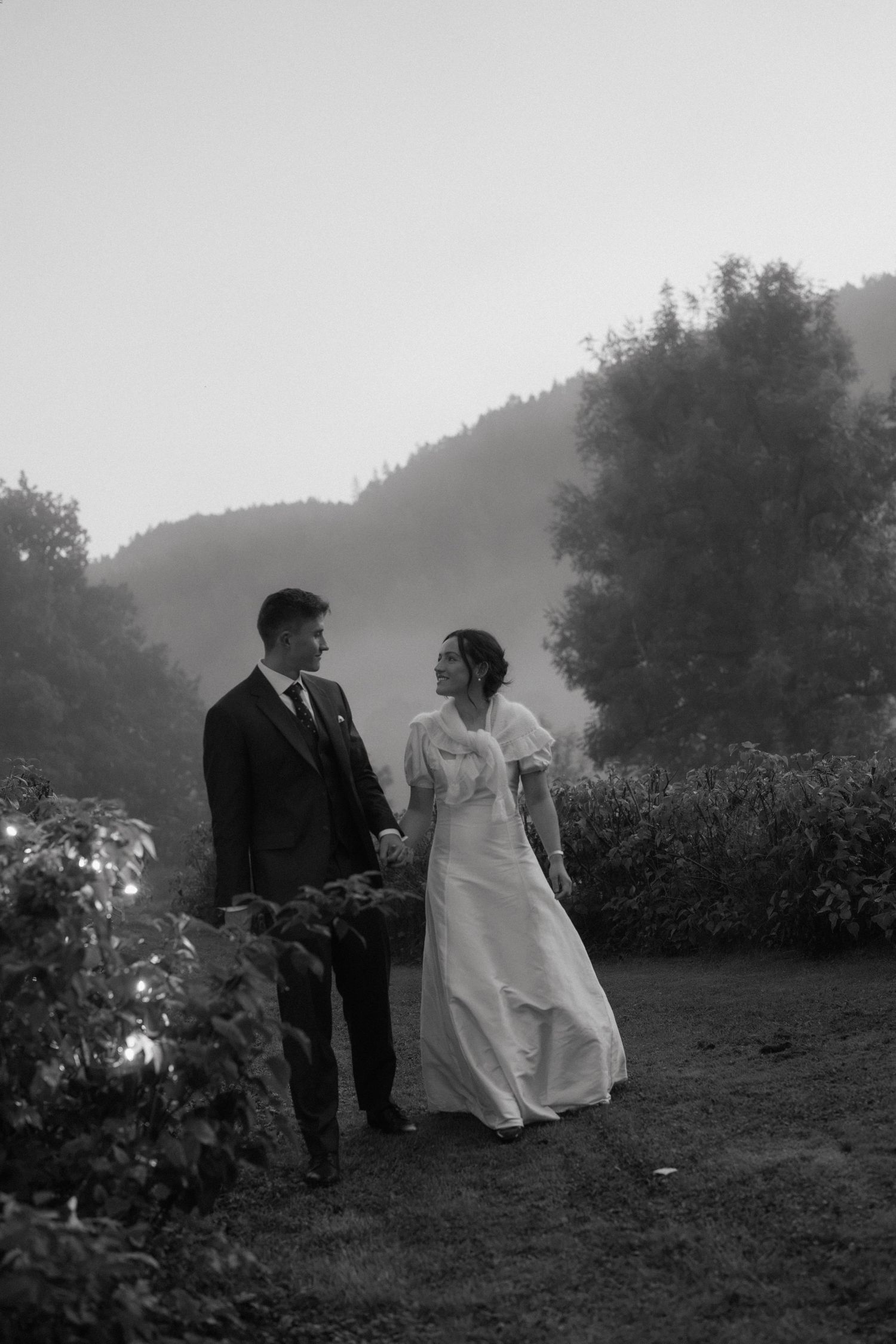 Black and white photo of bride and groom holding hands in misty outdoor landscape.