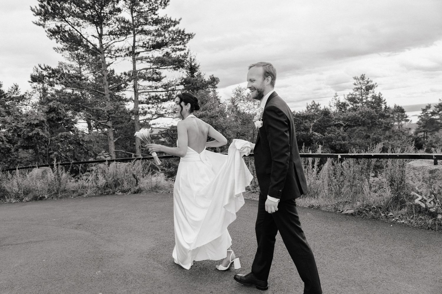 Black and white photo of a bride and groom walking outdoors, groom holding bride's dress train.
