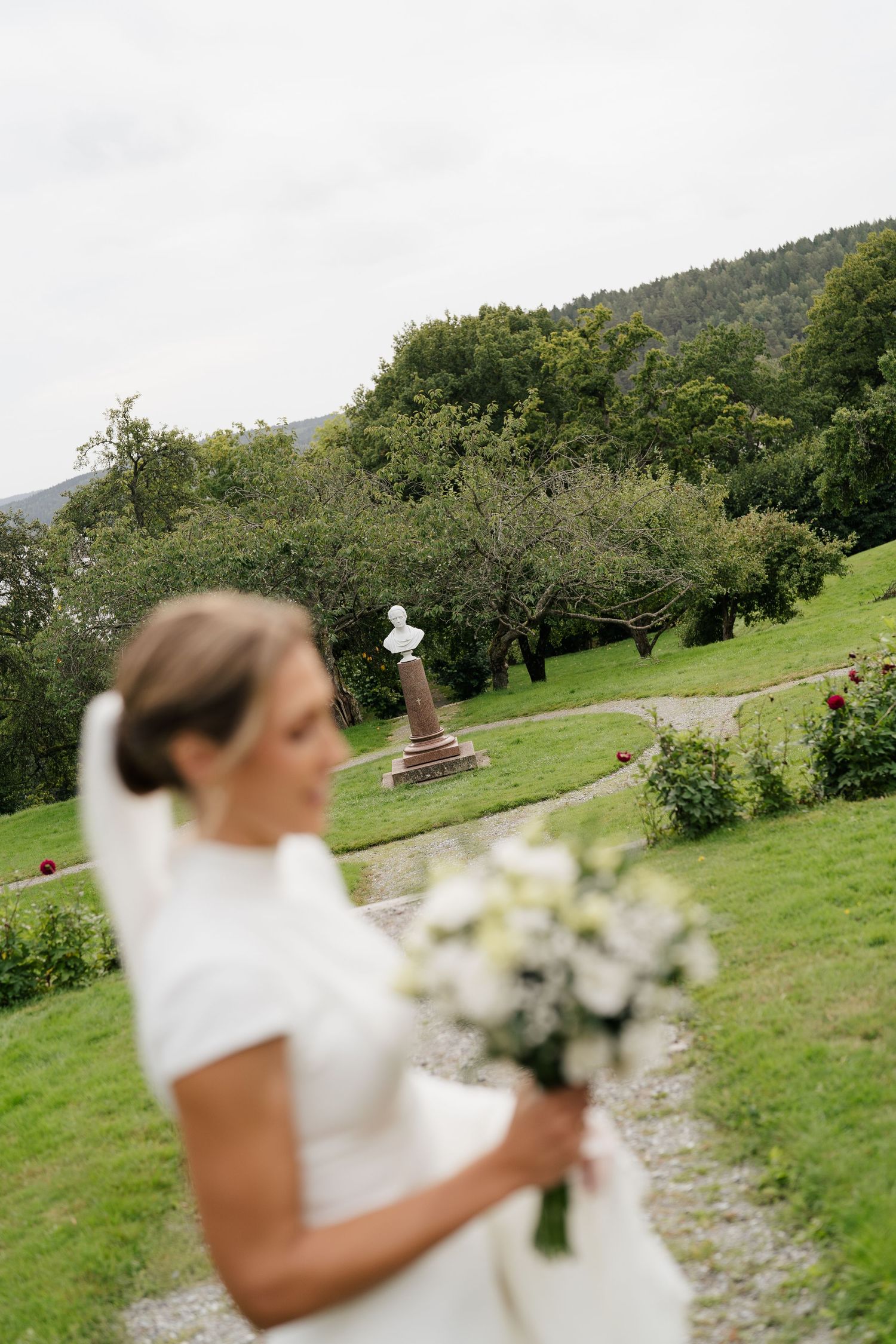 Elegant bride in white gown holding a white bouquet in a lush green garden with a classical stone monument.