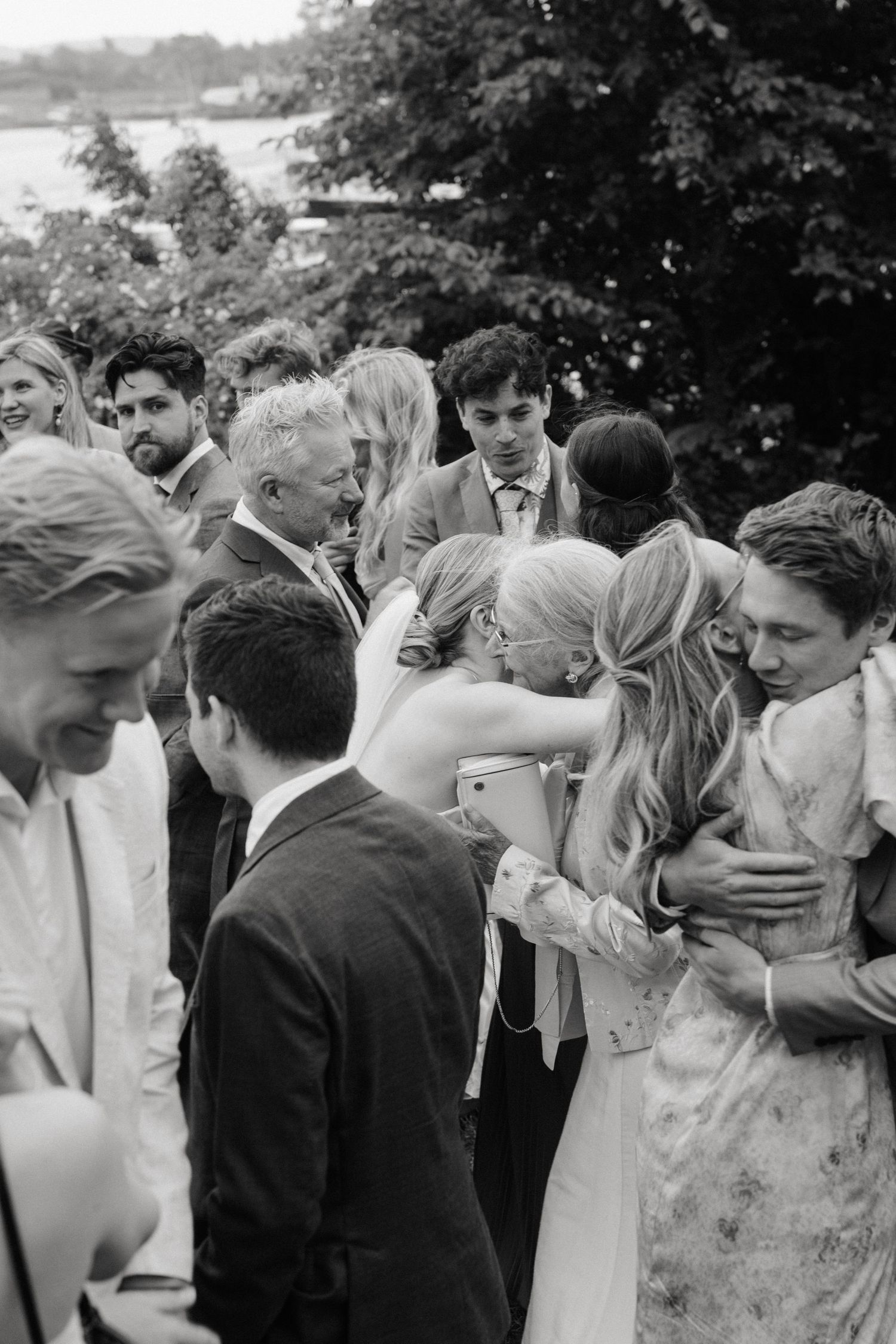 Black and white wedding photo of bride hugging guests outdoors surrounded by elegantly dressed attendees.