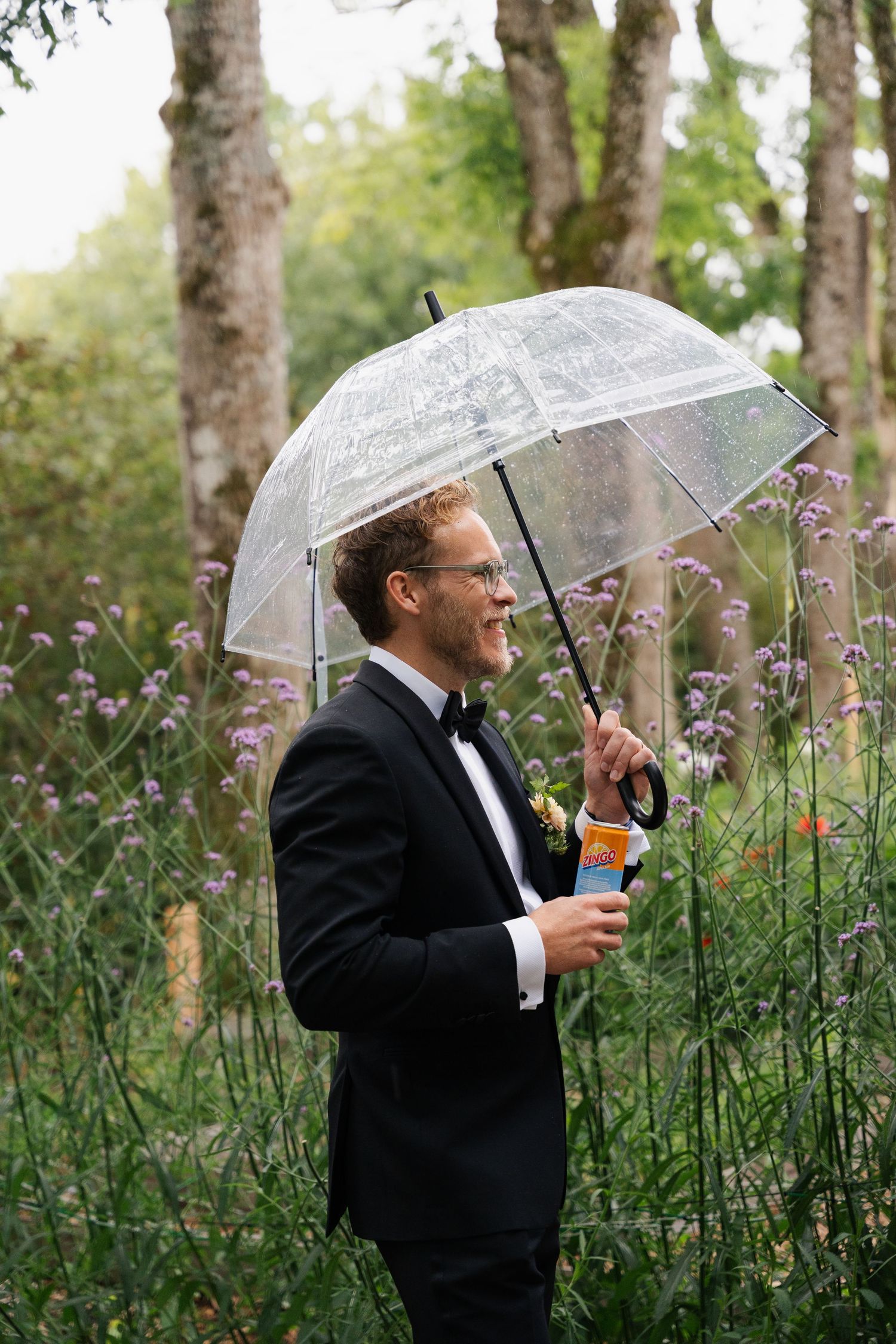 Dapper groom in a tuxedo holding a clear umbrella among purple wildflowers in a garden setting.