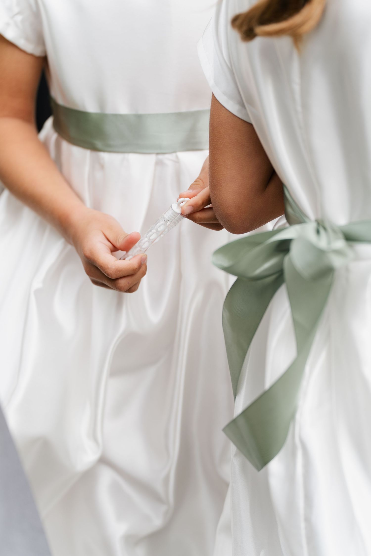 Two young flower girls in white dresses with sage green sashes fasten each others buttons.