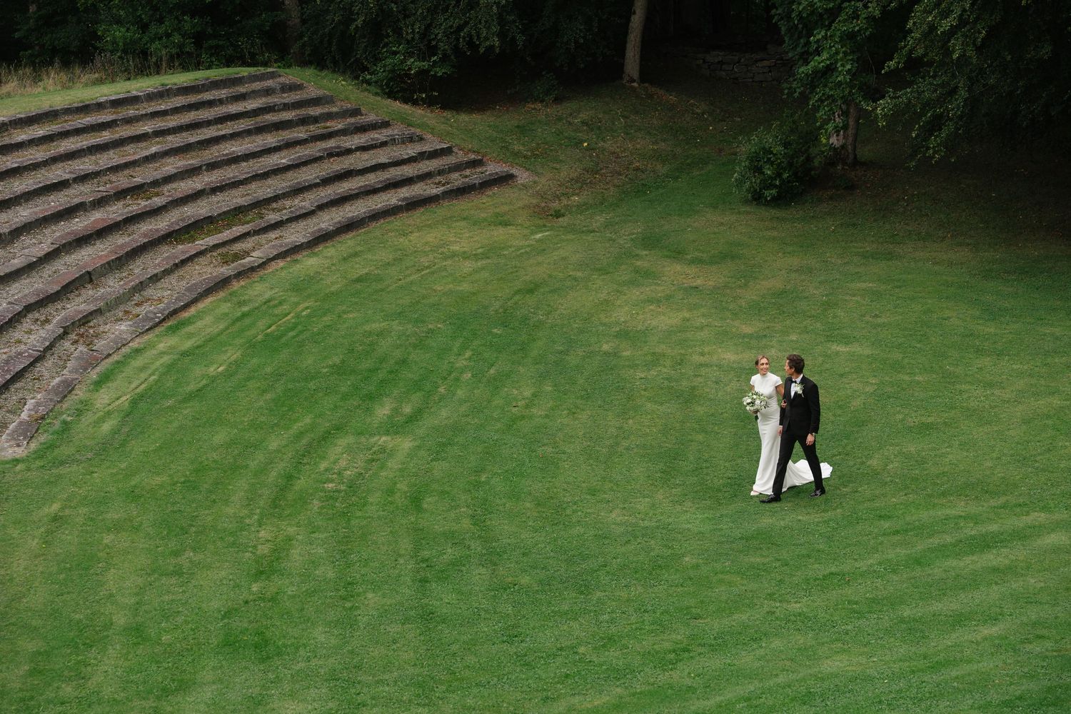 Aerial view of bride and groom walking across manicured green lawn near stone amphitheater steps.