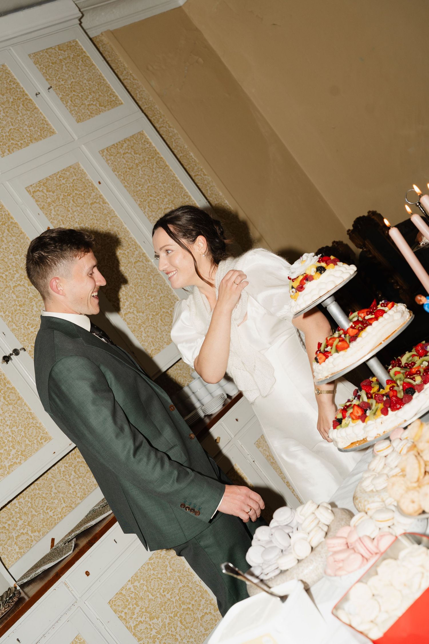 Bride and groom smile together beside a decorated multi-tiered wedding cake with fresh fruit.