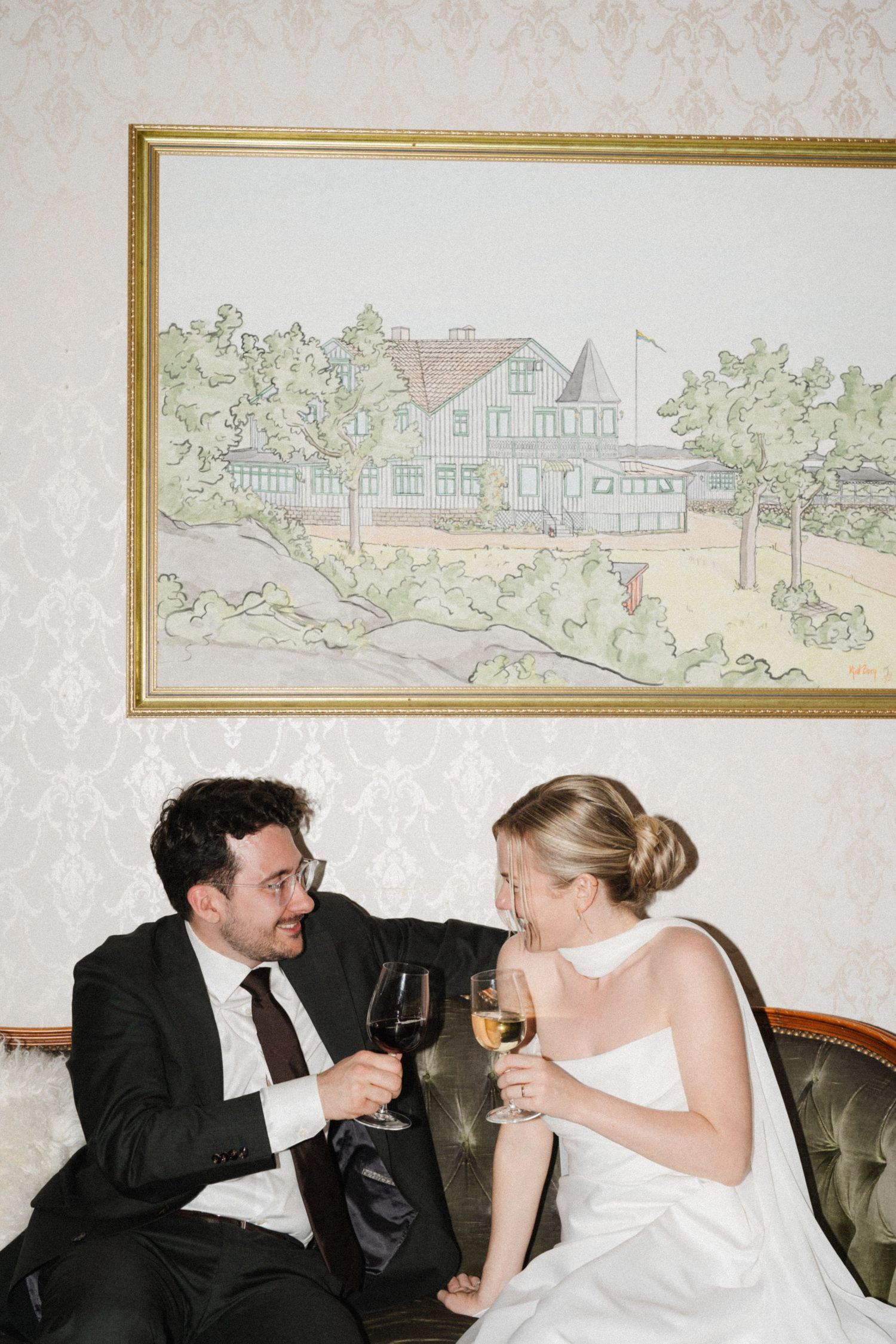 Bride and groom toast with wine glasses while seated on a vintage sofa beneath a painting.