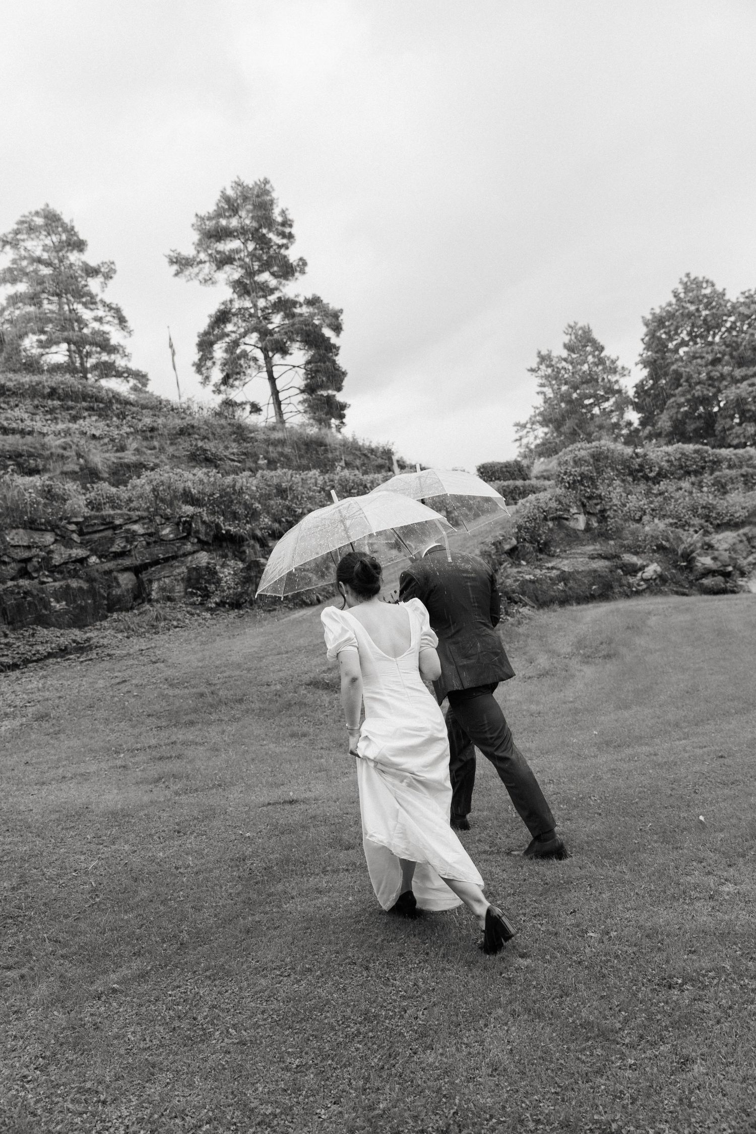 Black and white photo of bride and groom walking with clear umbrella on rainy wedding day.
