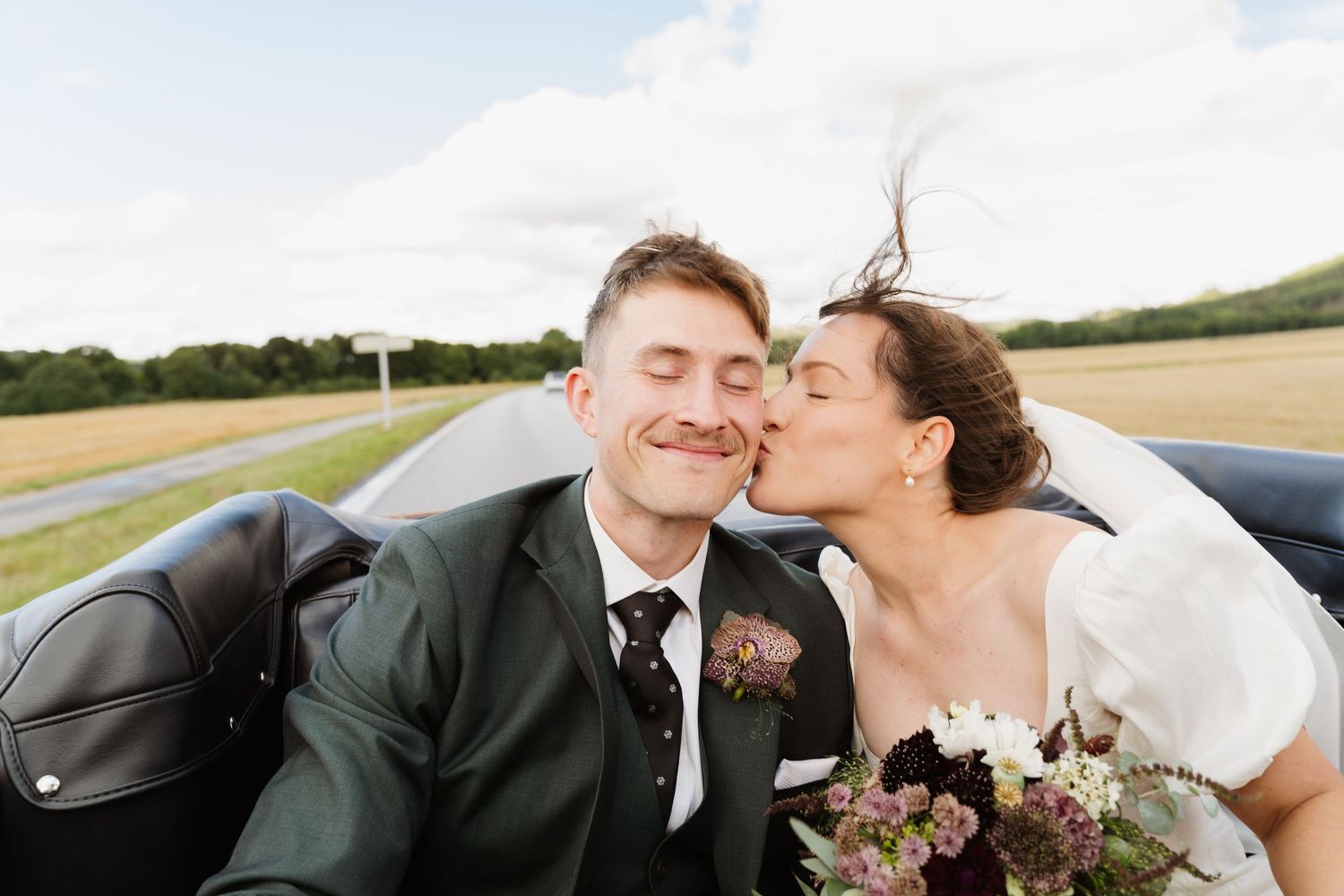 Bride kisses groom on cheek in convertible car with bouquet, countryside backdrop.