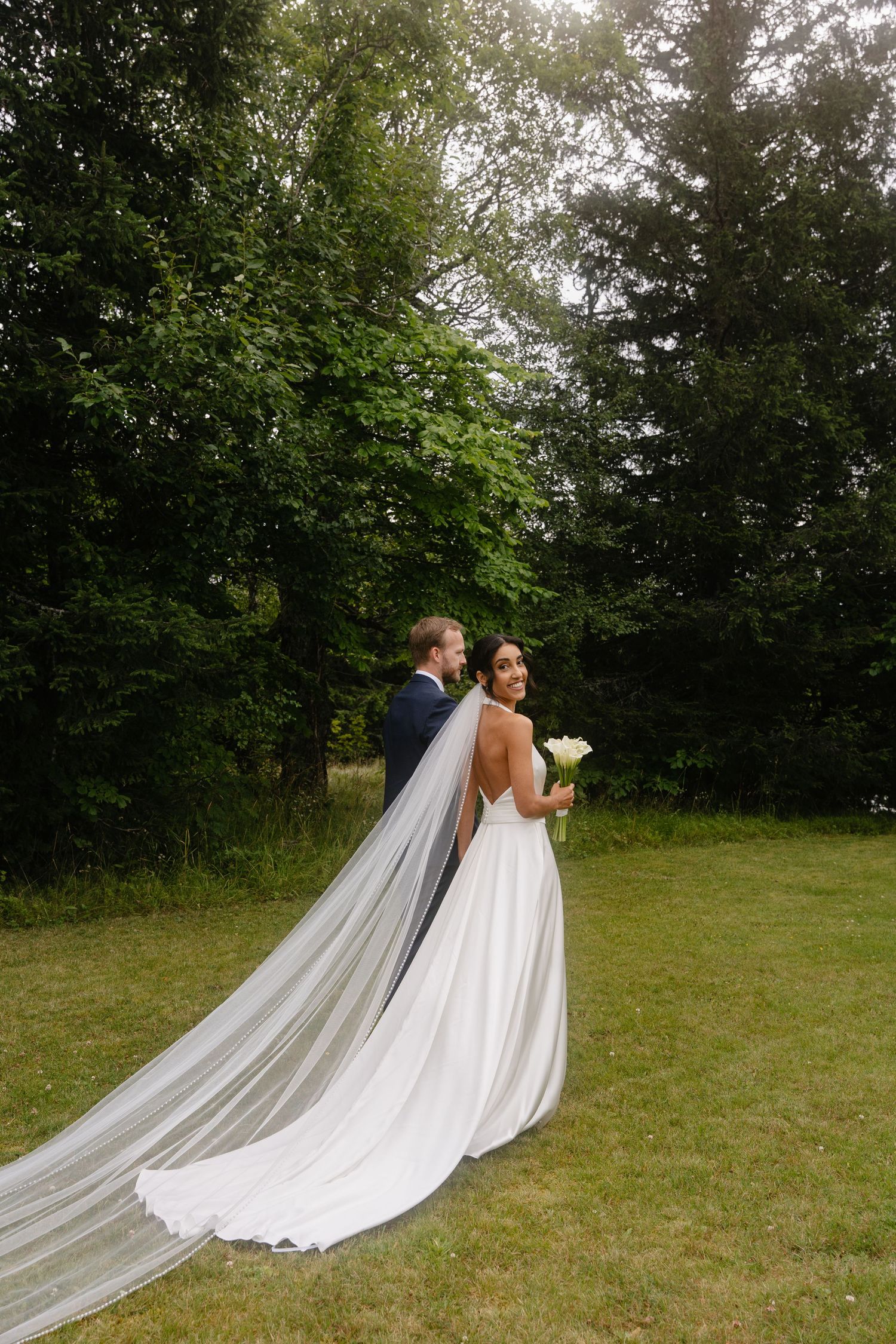 Bride in a white gown with long veil holds bouquet beside groom in navy suit, standing on green lawn near trees.