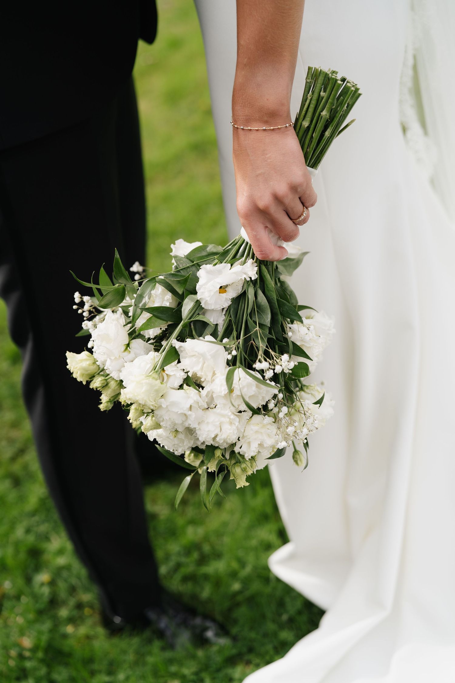 Bride in white gown holds a lush white floral bouquet beside groom on green grass.