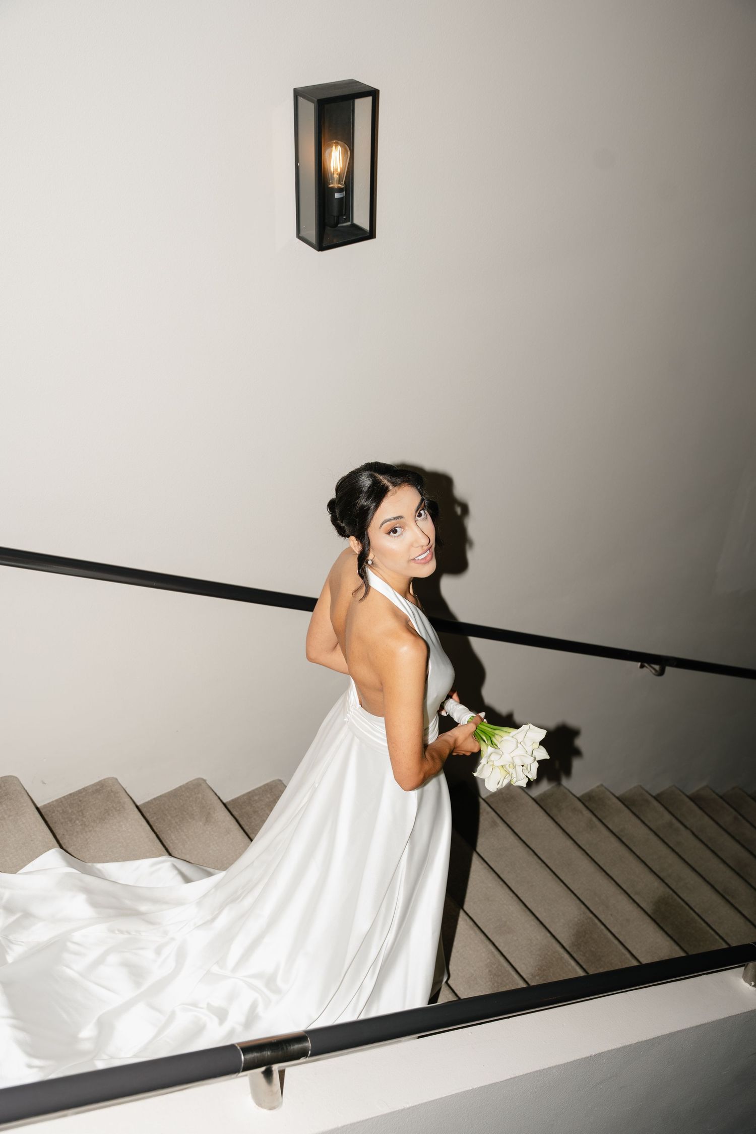 Elegant bride in a white halter gown holds a bouquet while posing on stairs with a wall sconce above.