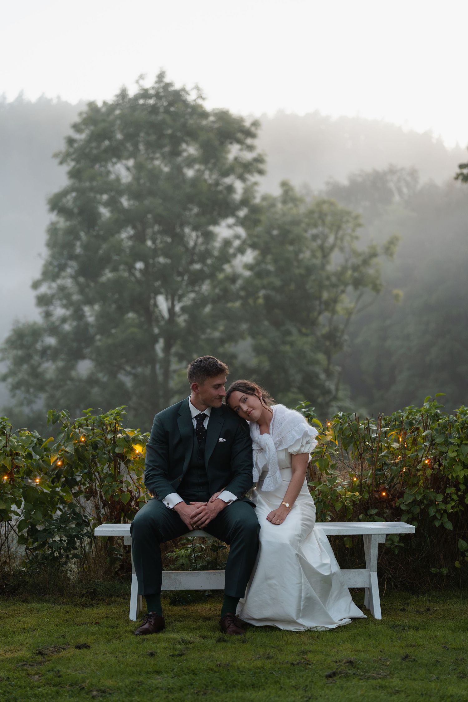 Bride and groom sitting on white bench in misty foggy garden at dusk during wedding.