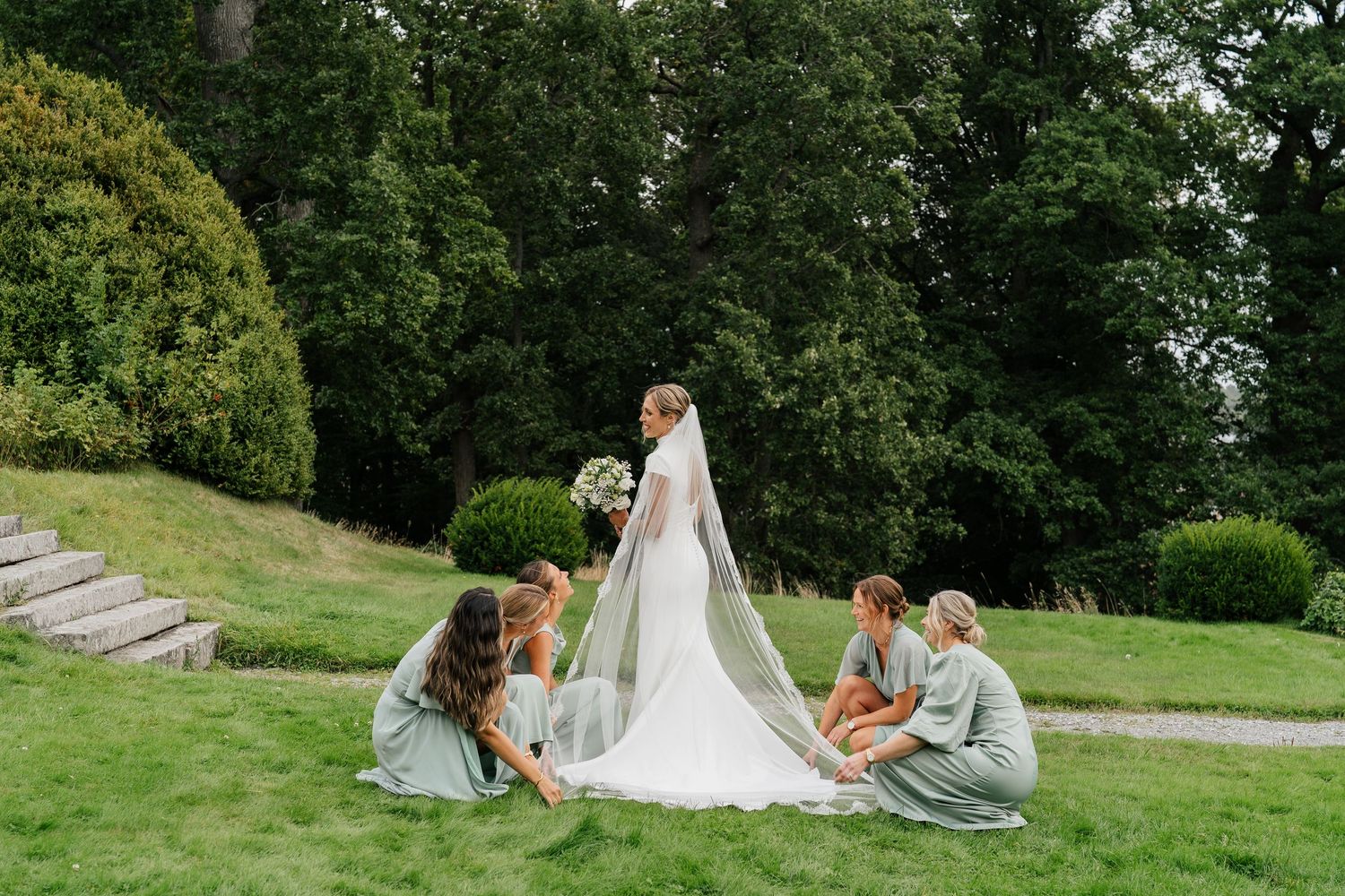 Bridesmaids in sage green dresses arrange bride's cathedral veil on lush garden lawn.